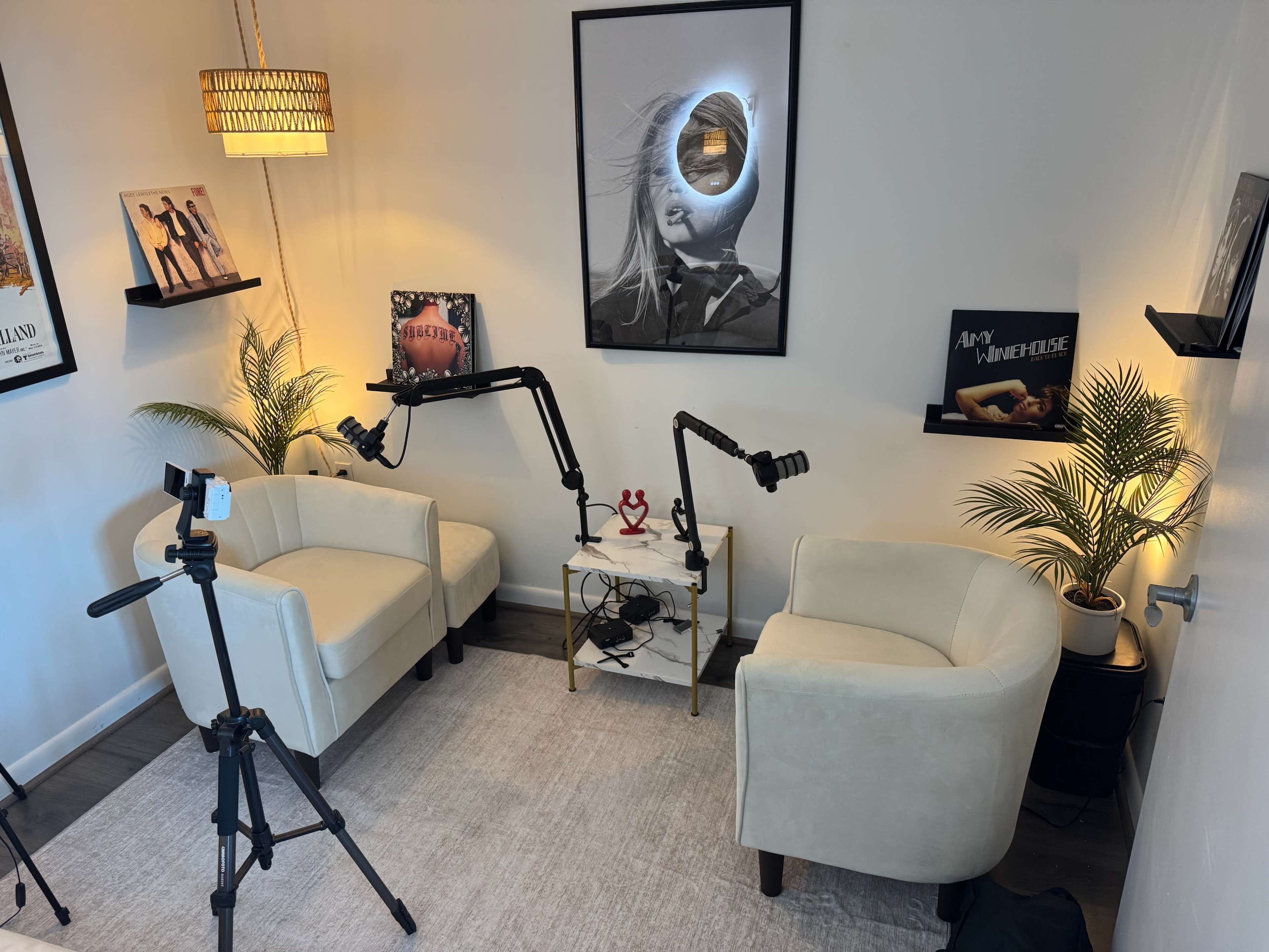 The image shows a small, modern room with two light-colored chairs, a small table with microphones mounted on arms, and framed artwork on the walls.