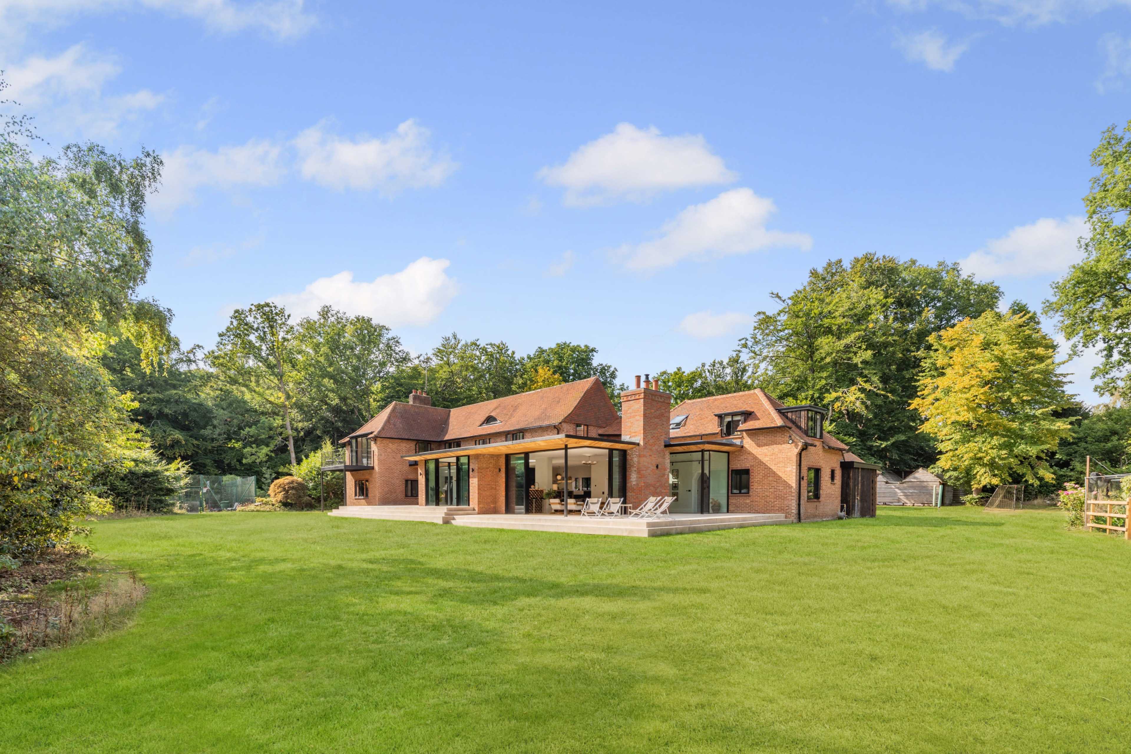 The image shows a brick house with large glass doors and a patio, set in a grassy area surrounded by trees.