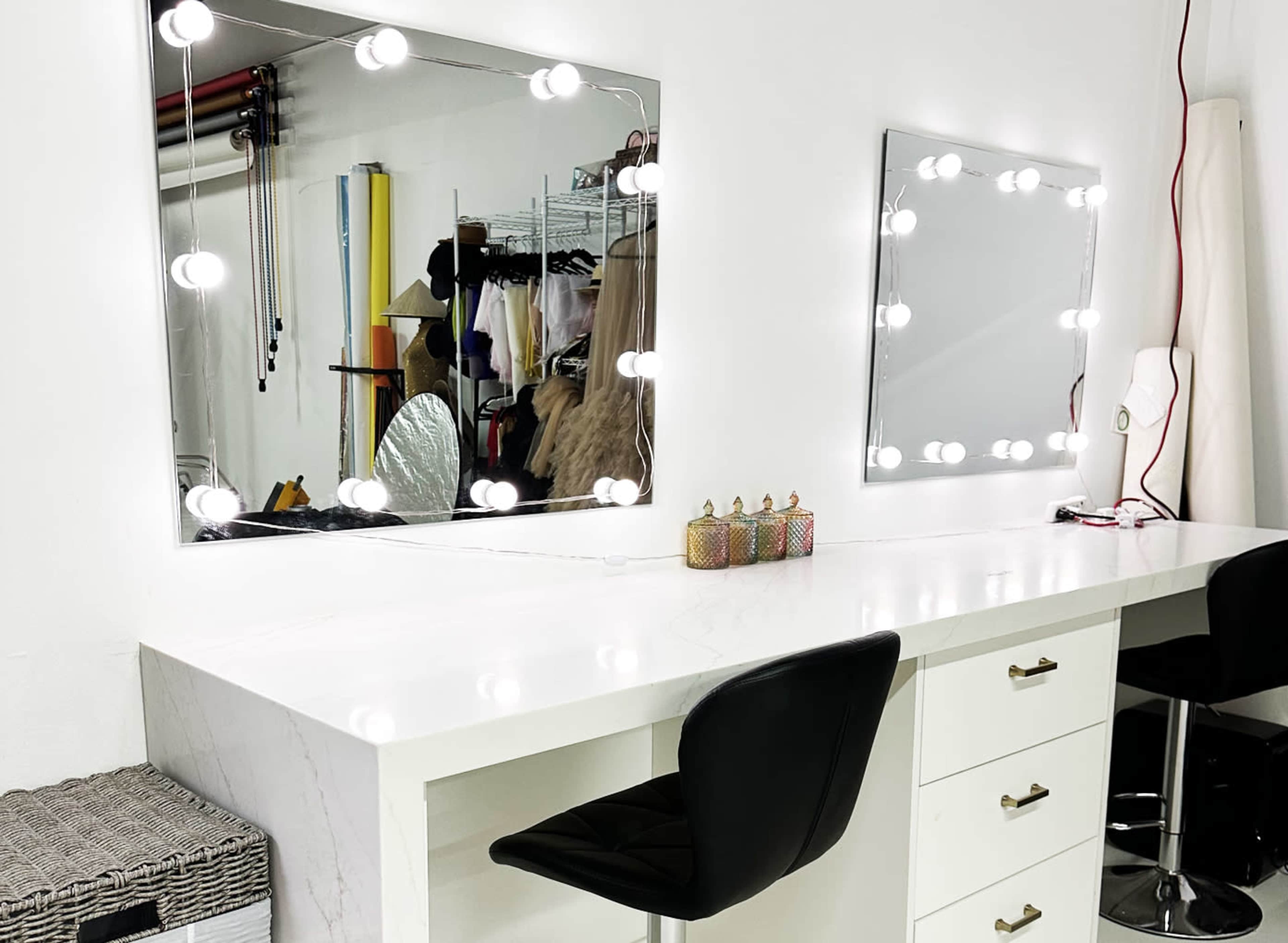 The image shows a bright, modern makeup station featuring two mirrors with light bulbs, a white marble countertop, and black chairs.