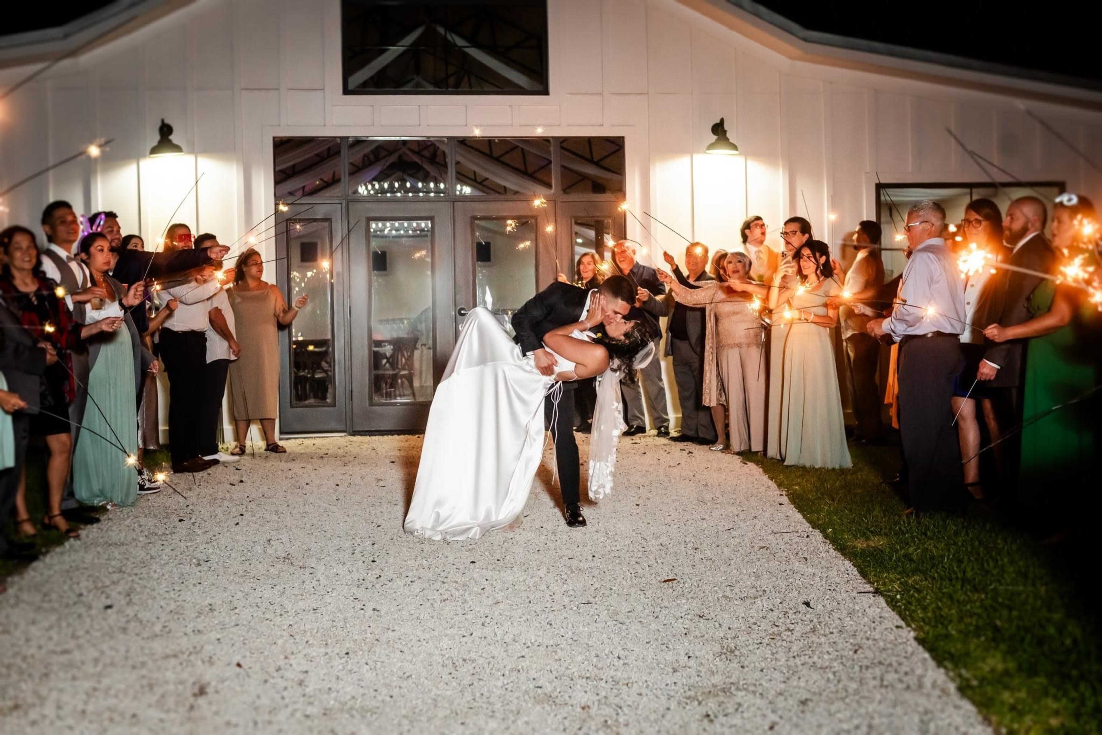A couple kisses as they are surrounded by wedding guests holding sparklers in a garden setting at night.