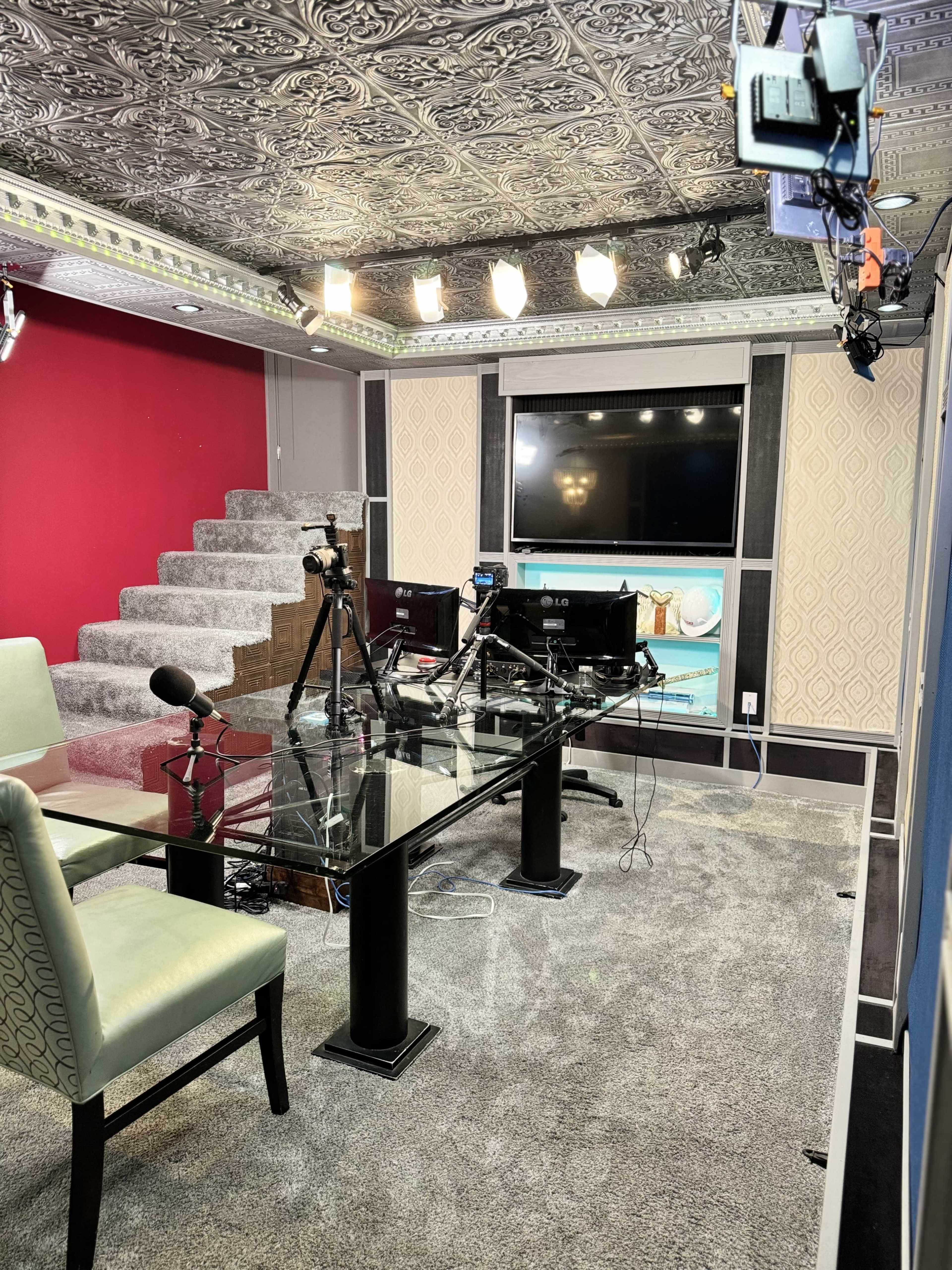 The image shows a modern recording studio with a glass table, two green chairs, multiple cameras and microphones, and a large screen on the wall.