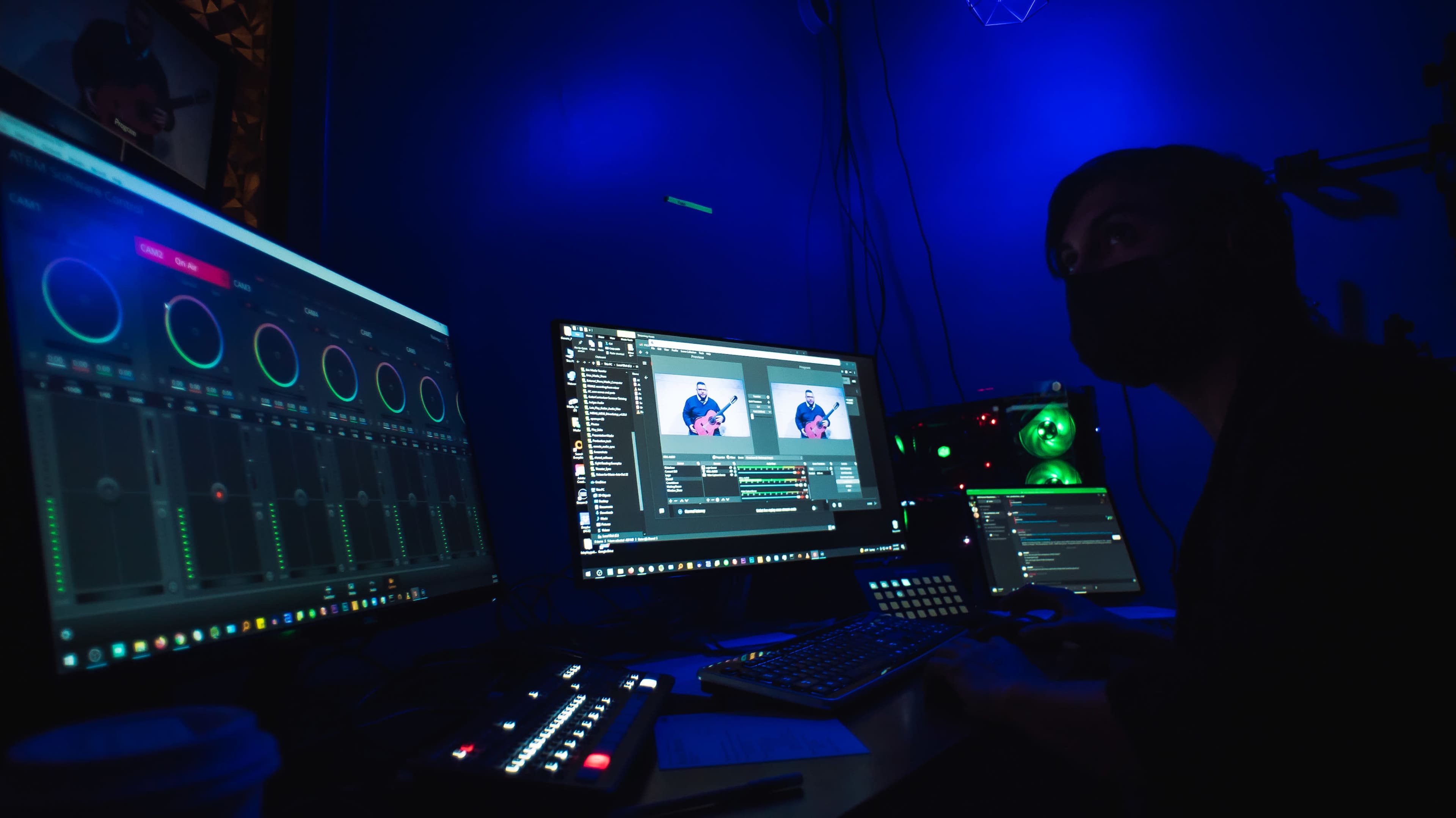 A person with a mask is seated at a desk in a dimly lit room, working on multiple computer monitors displaying editing software and audio controls.