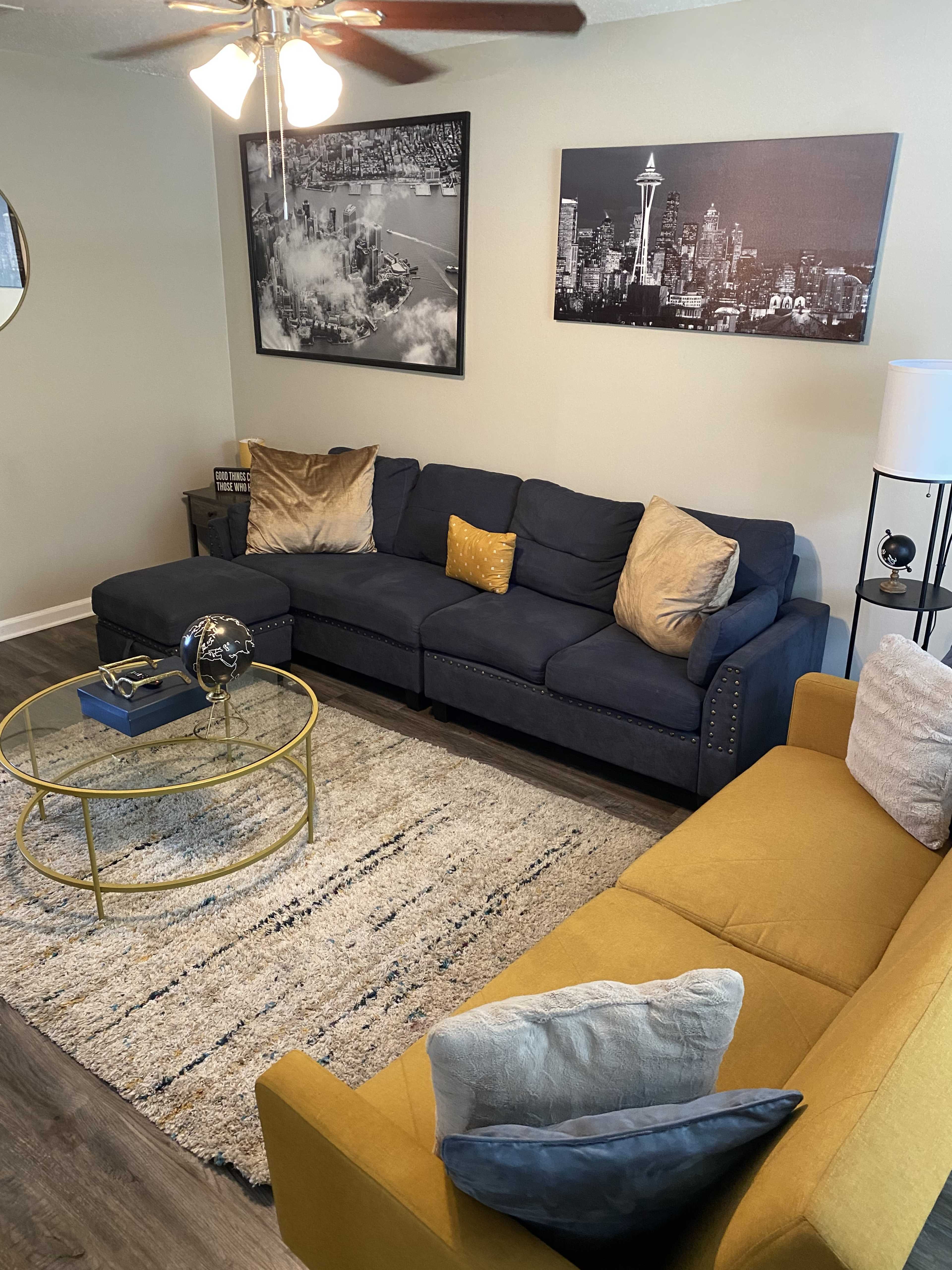 A living room with a navy blue sectional sofa, a yellow accent sofa, a round coffee table, and two framed black-and-white cityscape photos on the wall.