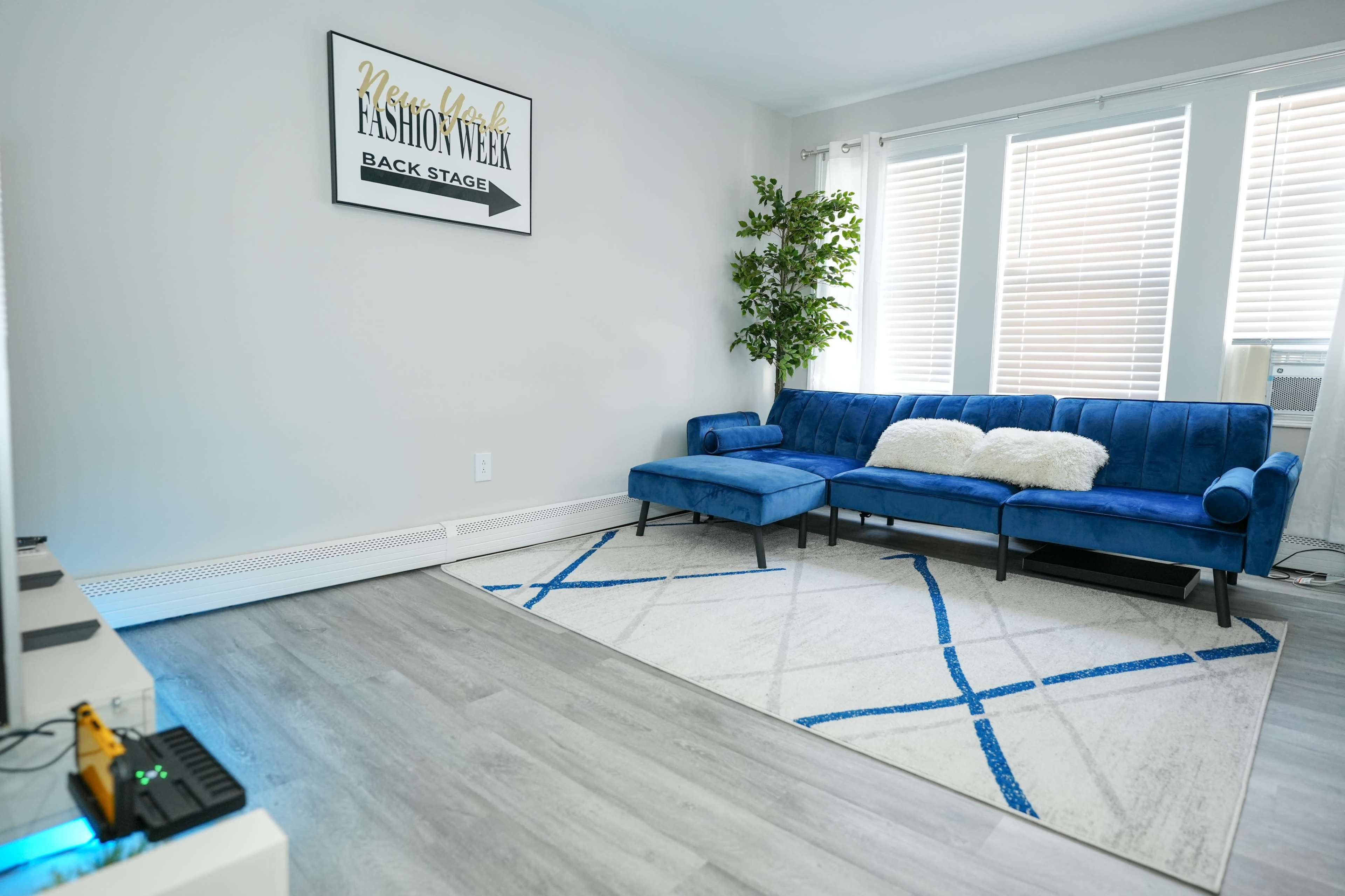 A spacious living room with a blue sectional sofa, a patterned rug, and a framed poster on the wall, featuring "Fashion Week" text.