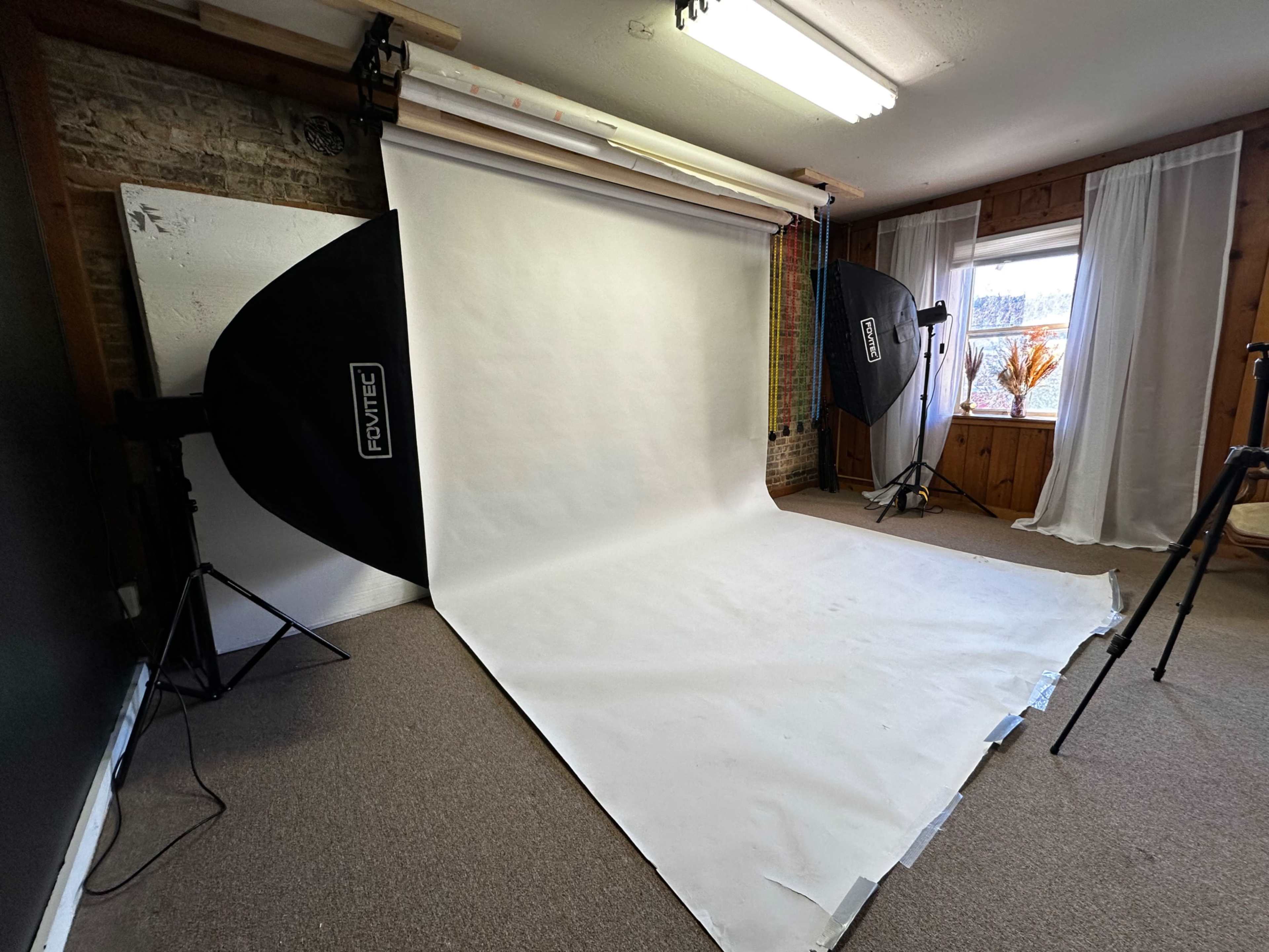 A photography studio with a white backdrop, softbox lights, and a window with sheer curtains.