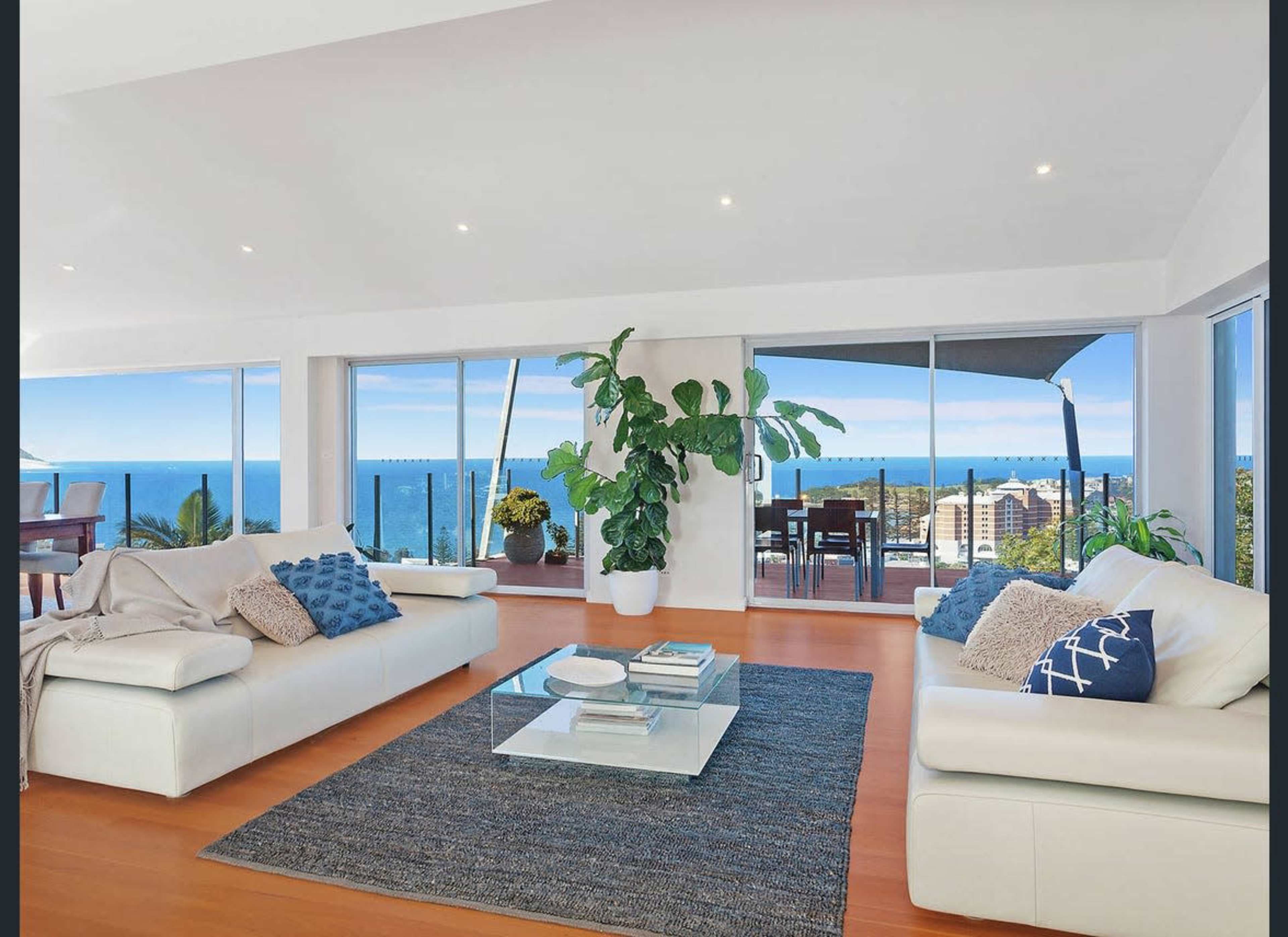 A modern living room with two white sofas, a glass coffee table, and a view of the ocean through large windows.