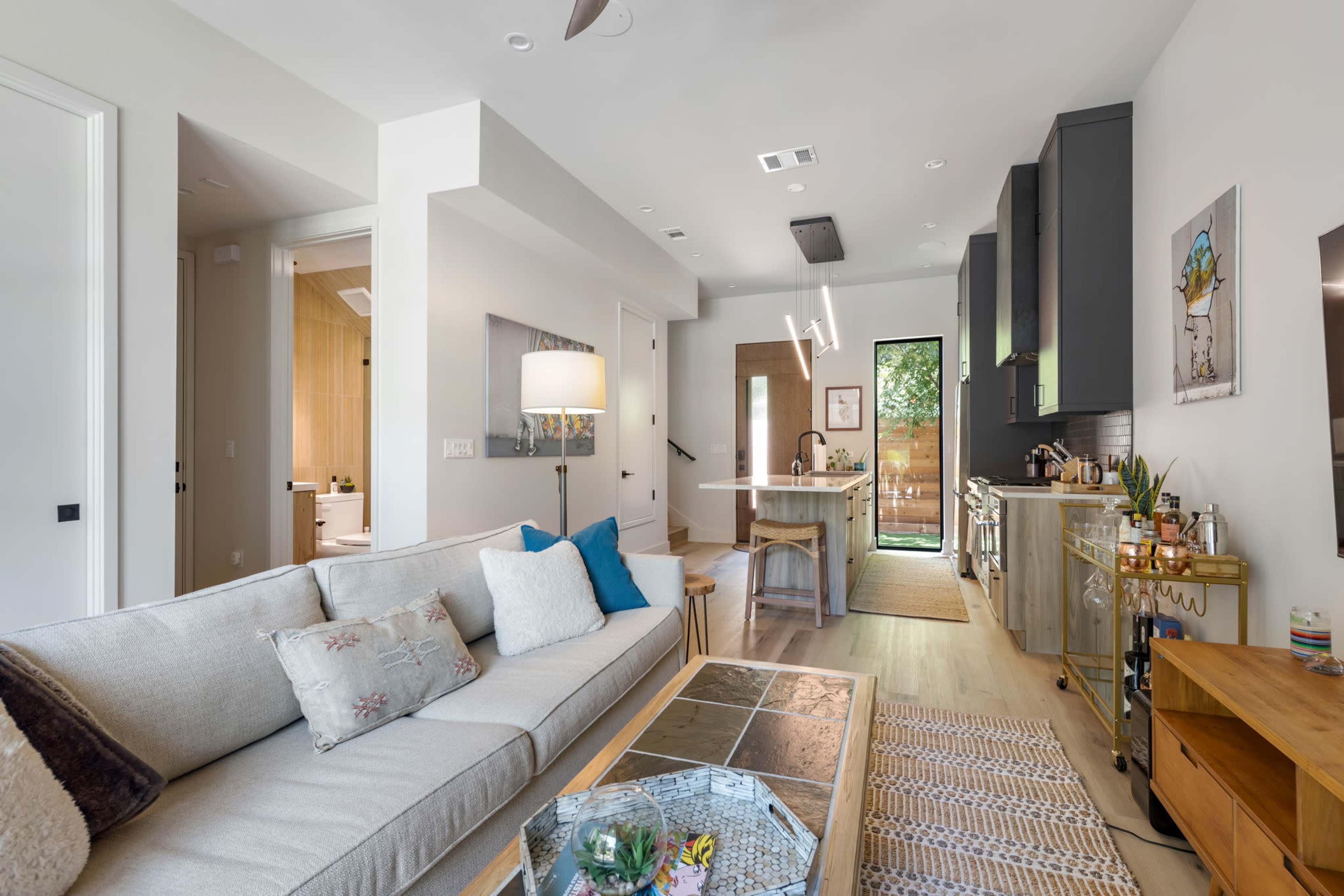 The image shows a modern, open-concept living space featuring a light-colored sofa, a wooden dining table, and a kitchen with dark cabinetry.