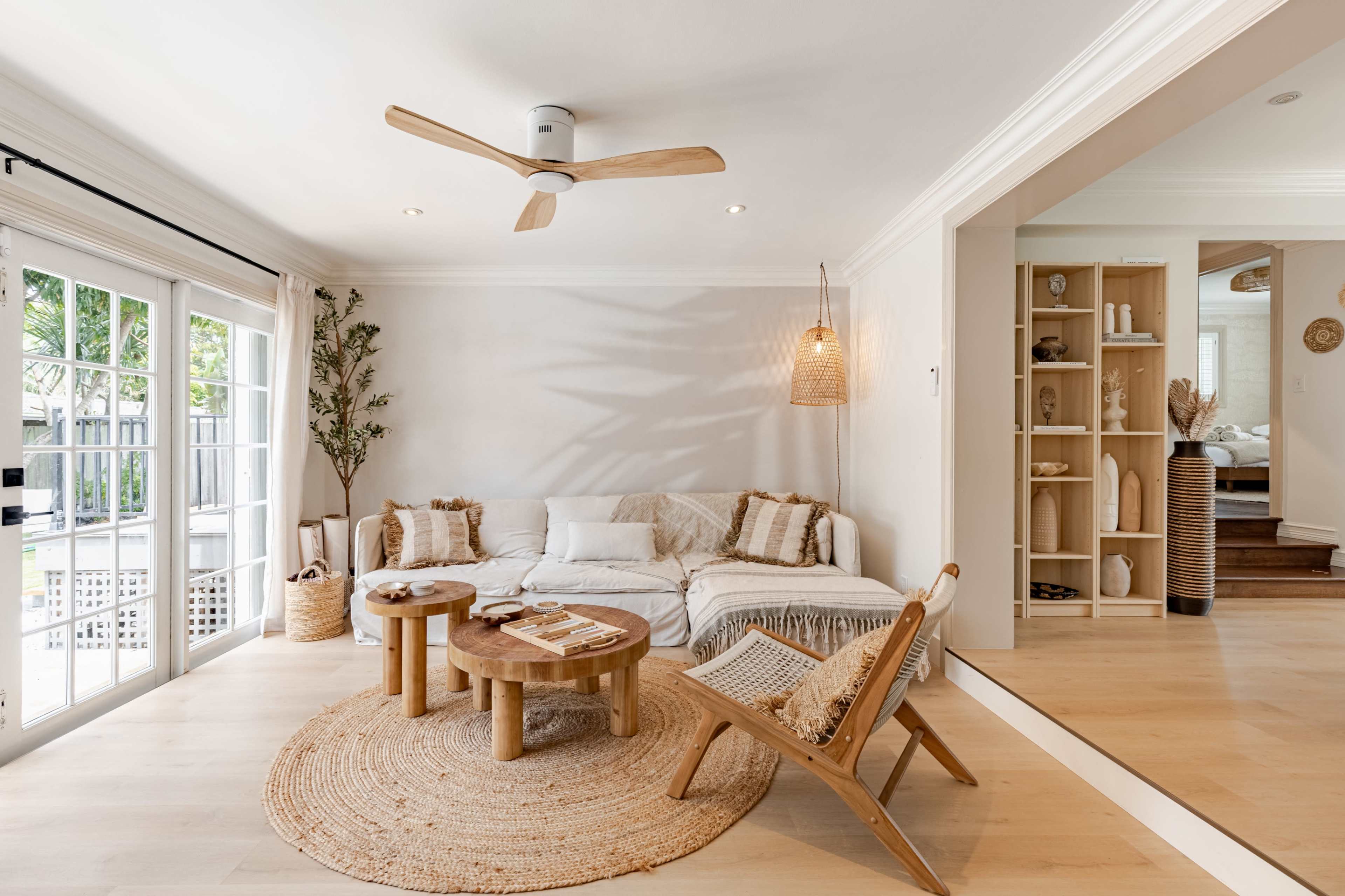 The living room features a light-colored sofa, two wooden tables, a natural fiber rug, and large windows that open to an outdoor space.