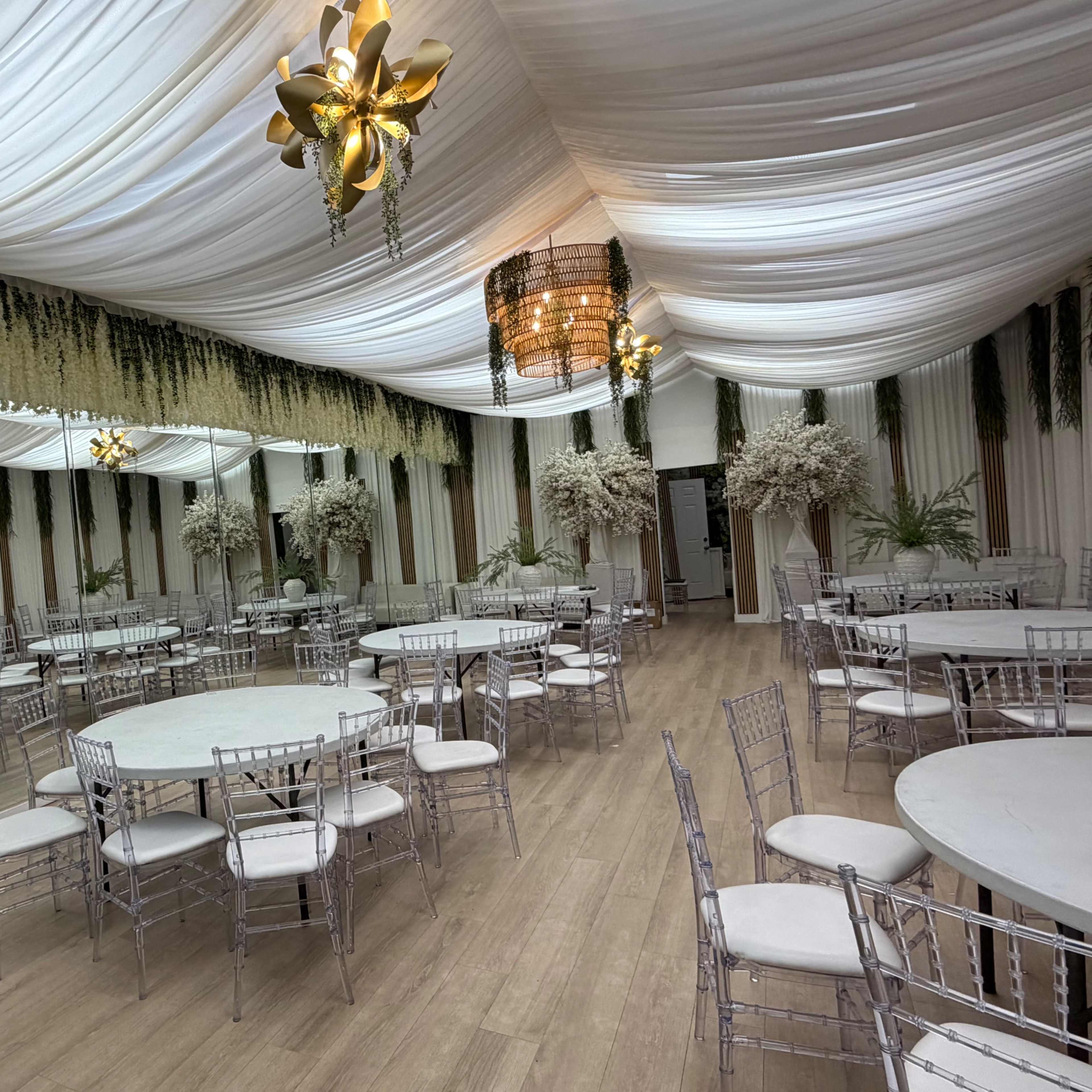 The image shows an elegantly decorated indoor event space with round tables, silver chiavari chairs, and floral arrangements under a draped ceiling.