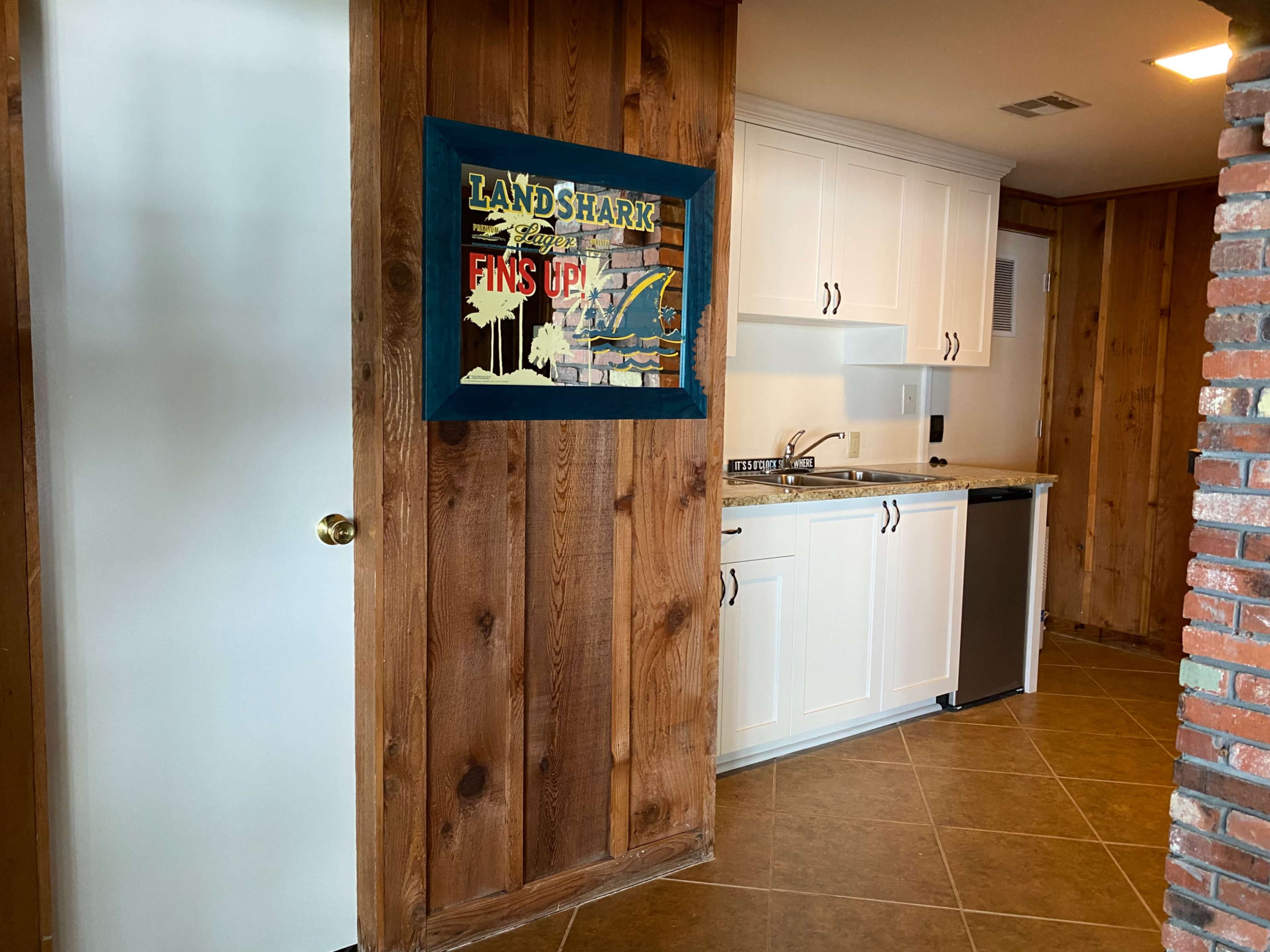 The image shows a kitchen area with white cabinetry, a brick accent wall, and a framed sign reading "LANDSHARK" hung on a wooden wall.