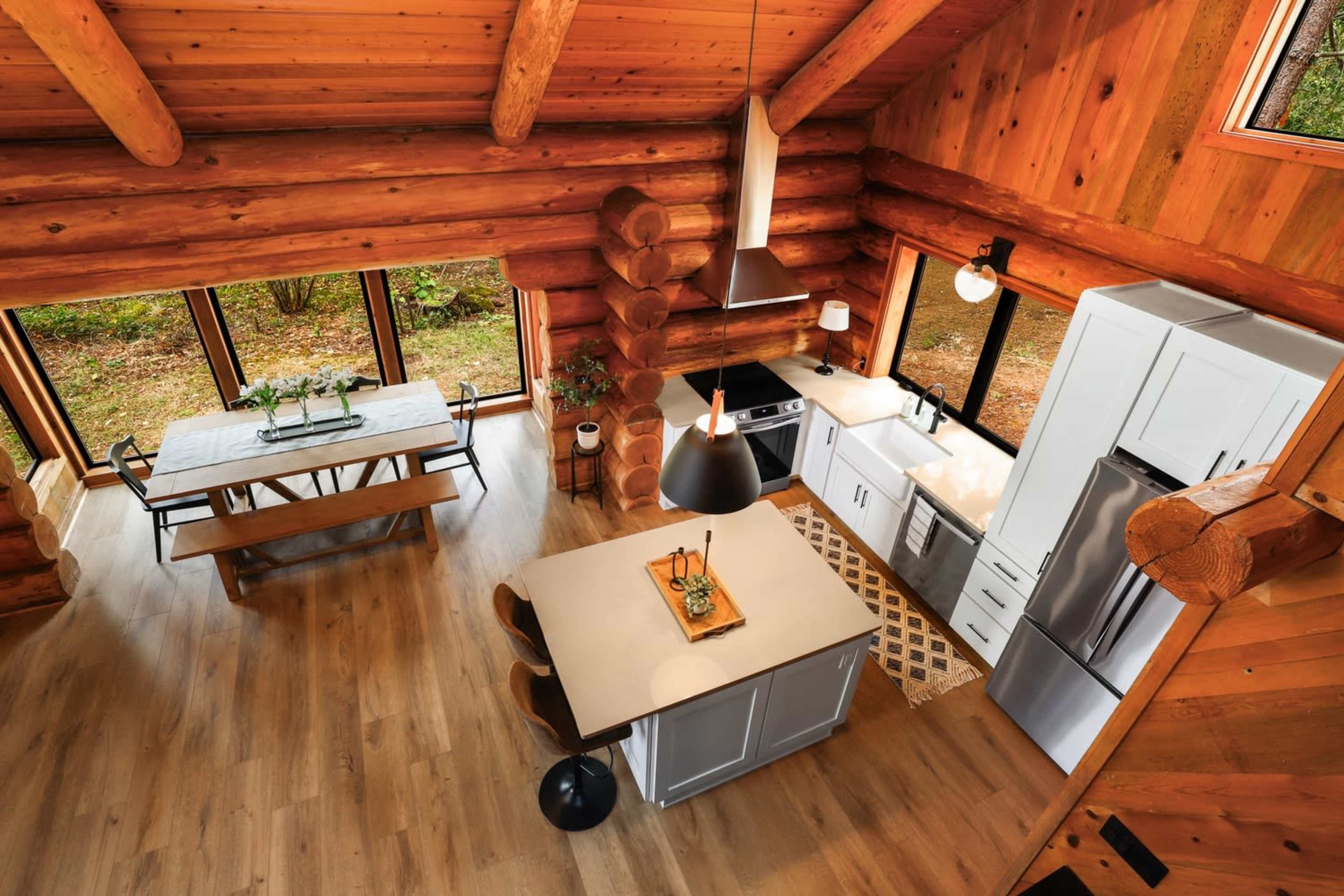 The image shows a spacious log cabin interior featuring a kitchen with modern appliances, a central island, and a dining area with a wooden table.