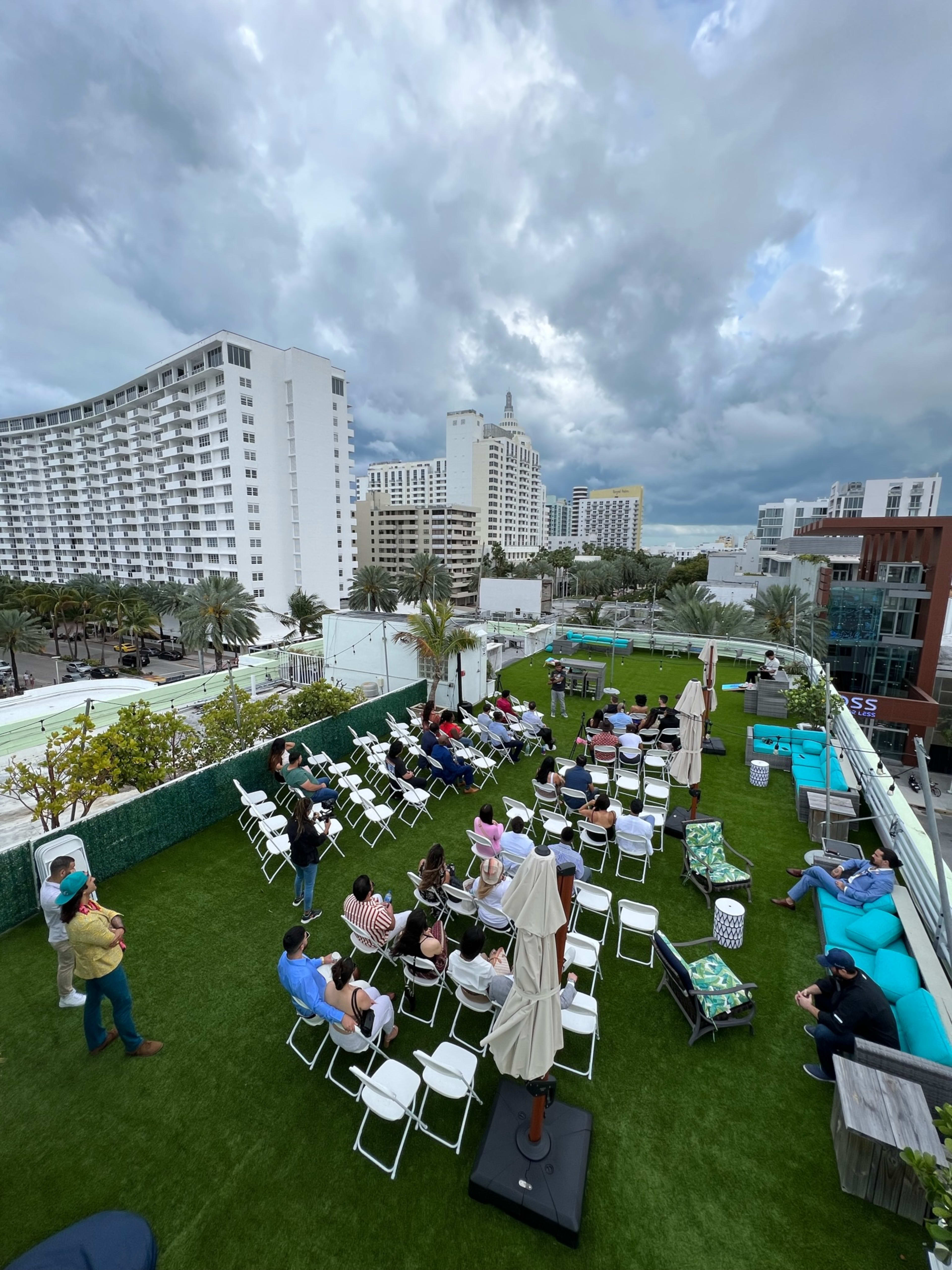A rooftop event featuring attendees seated in white chairs on a grass-like surface, surrounded by palm trees and high-rise buildings under a cloudy sky.