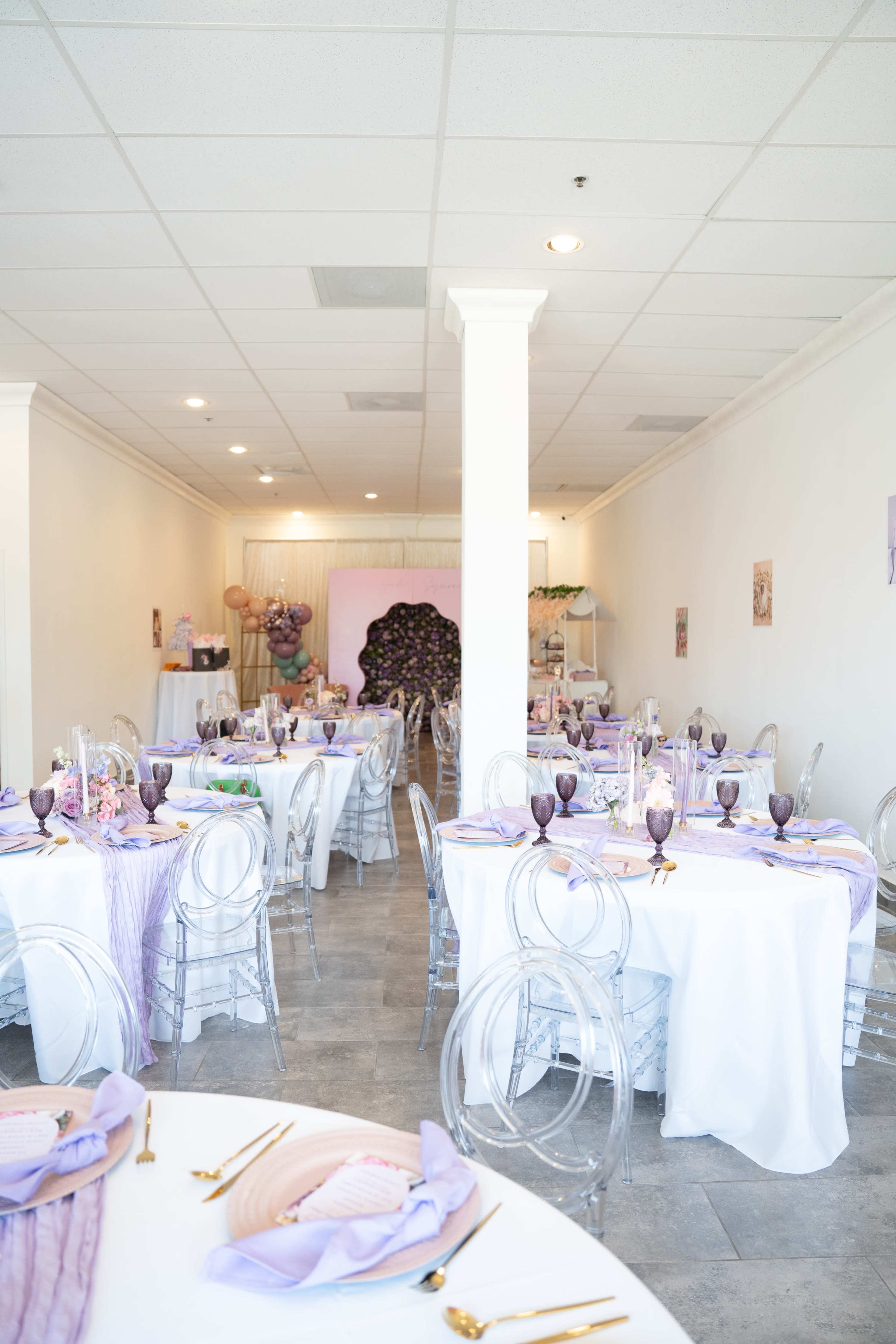 A spacious event venue is set up with several round tables covered in white linens and adorned with purple tableware, while a decorative backdrop featuring purple balloons is visible in the background.