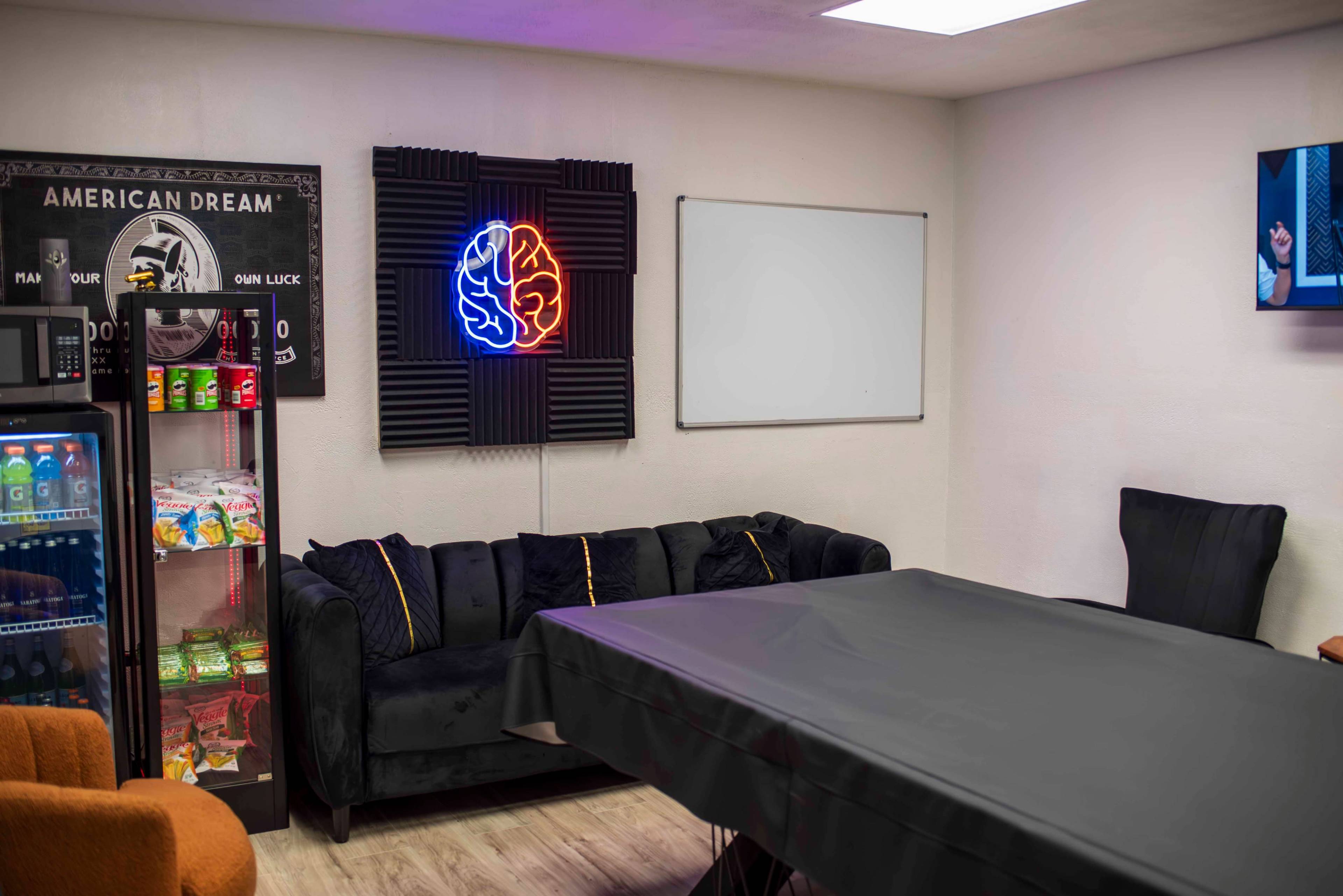 The image shows a cozy room with a black couch, a large table covered with a dark tablecloth, a neon brain display on the wall, and a refrigerated vending machine in the corner.