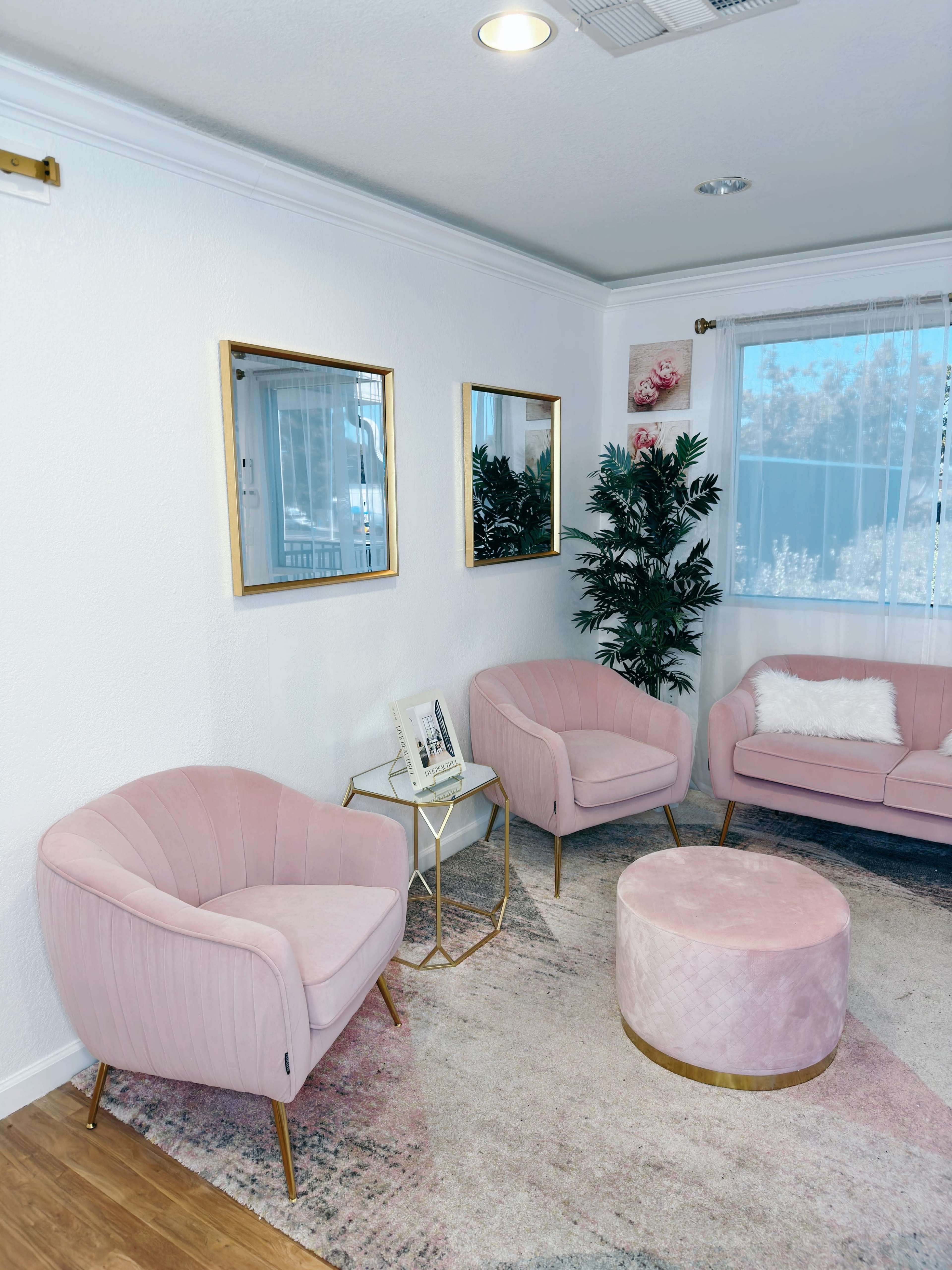 A modern living room with two pink armchairs, a pink ottoman, a small gold side table, and three mirrors on the wall.