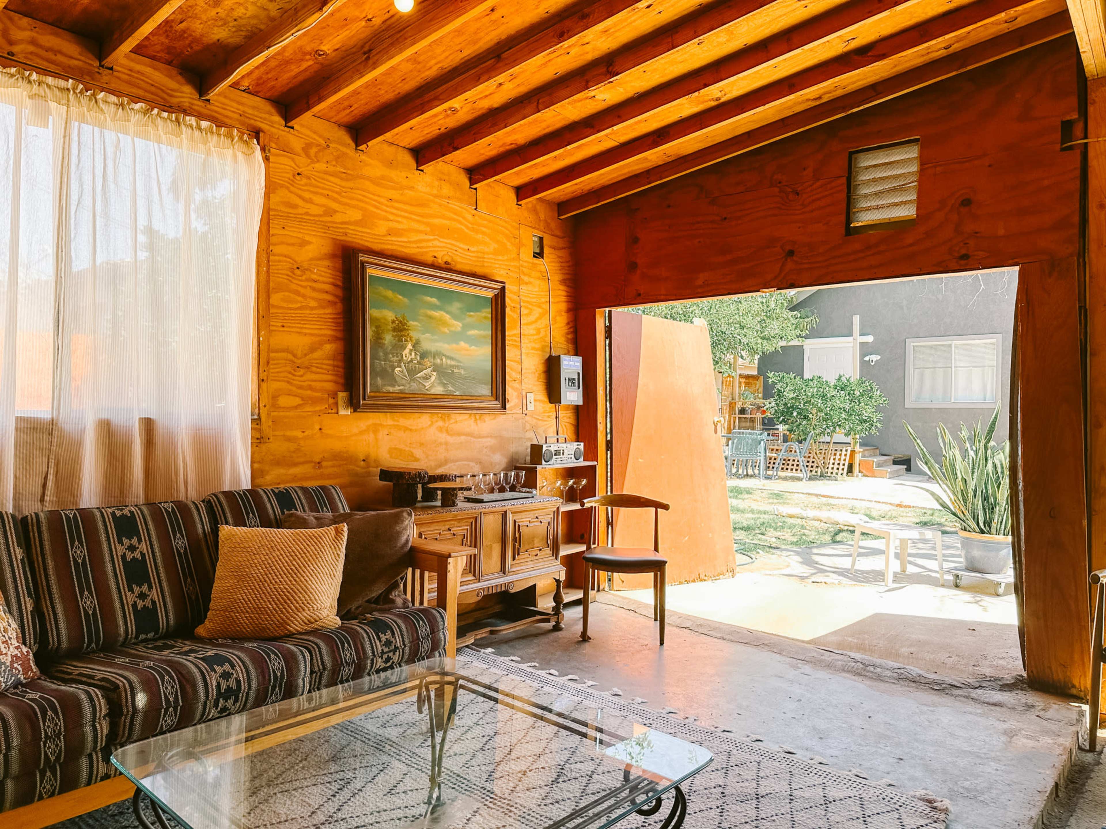 A cozy interior of a wooden room with a striped sofa, a glass coffee table, and an open door leading to a patio area with outdoor furniture.