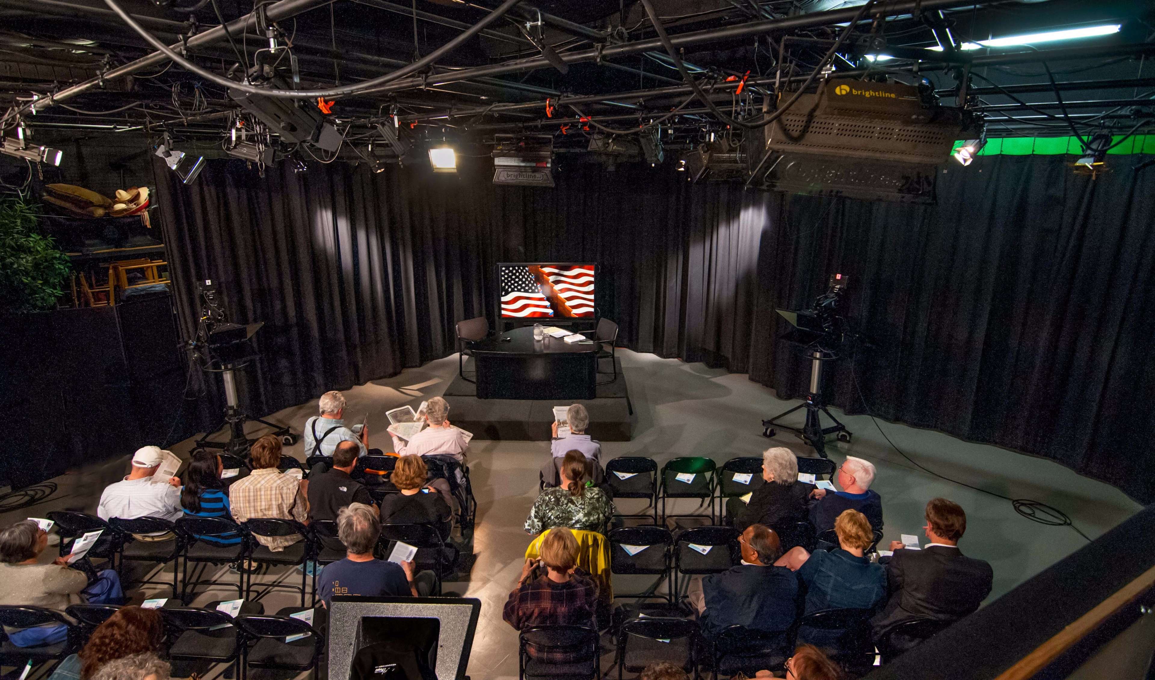 An indoor studio setup with a seated audience facing a presentation screen displaying American flags.