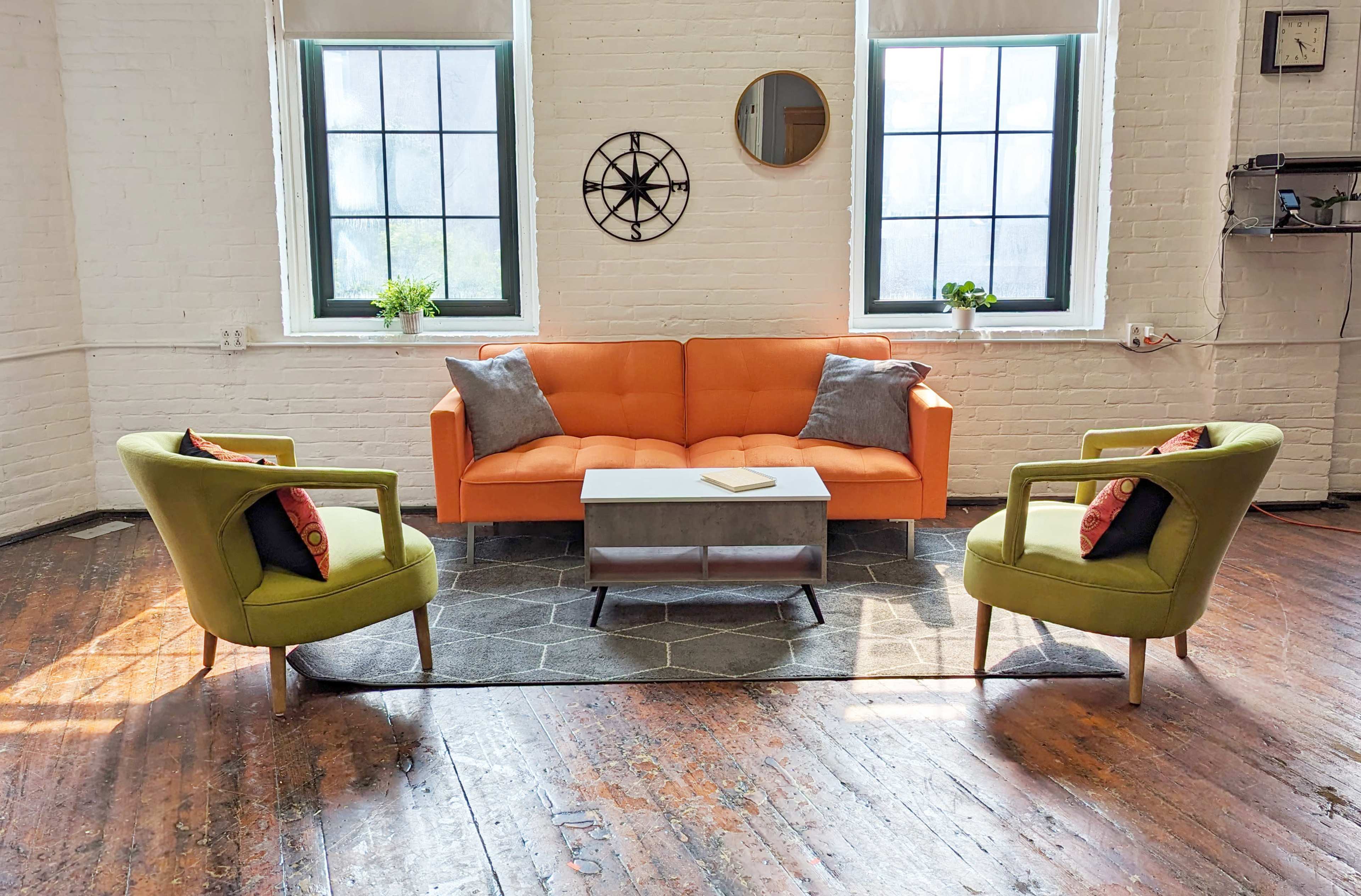A cozy living room with an orange sofa, two green armchairs, a coffee table on a hexagonal rug, and large windows letting in natural light.