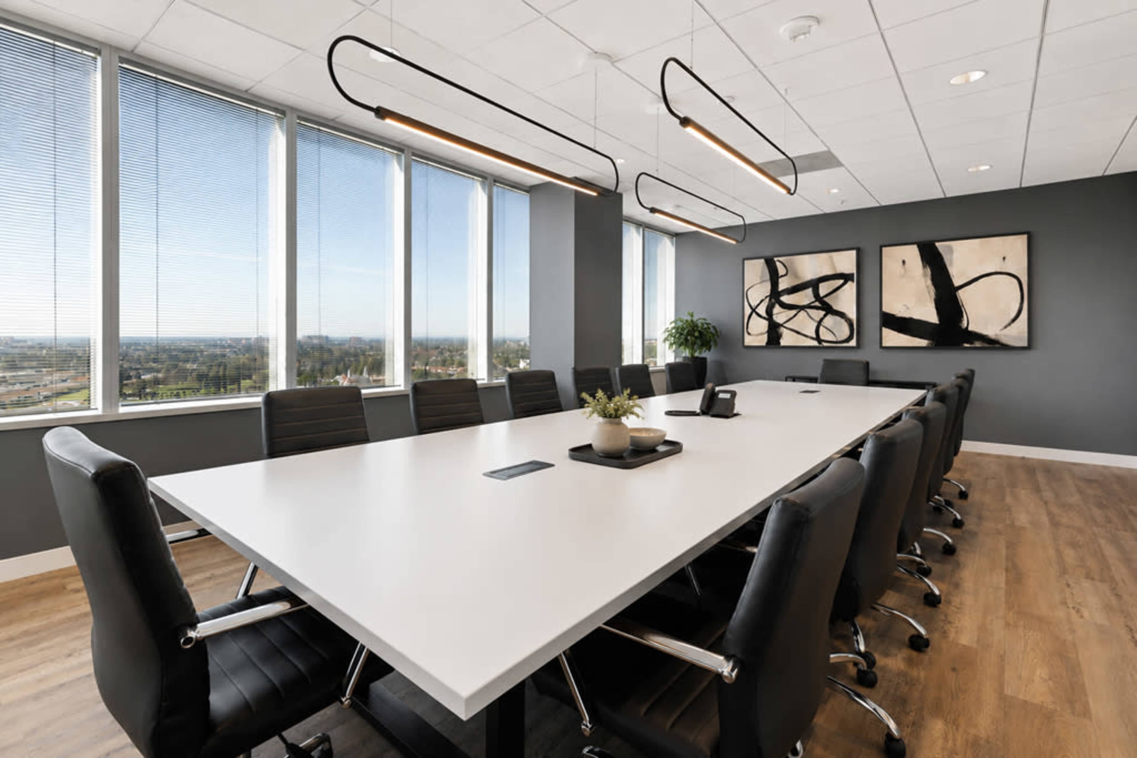 A large conference room features a long white table surrounded by black leather chairs, with floor-to-ceiling windows providing a view of the outside.