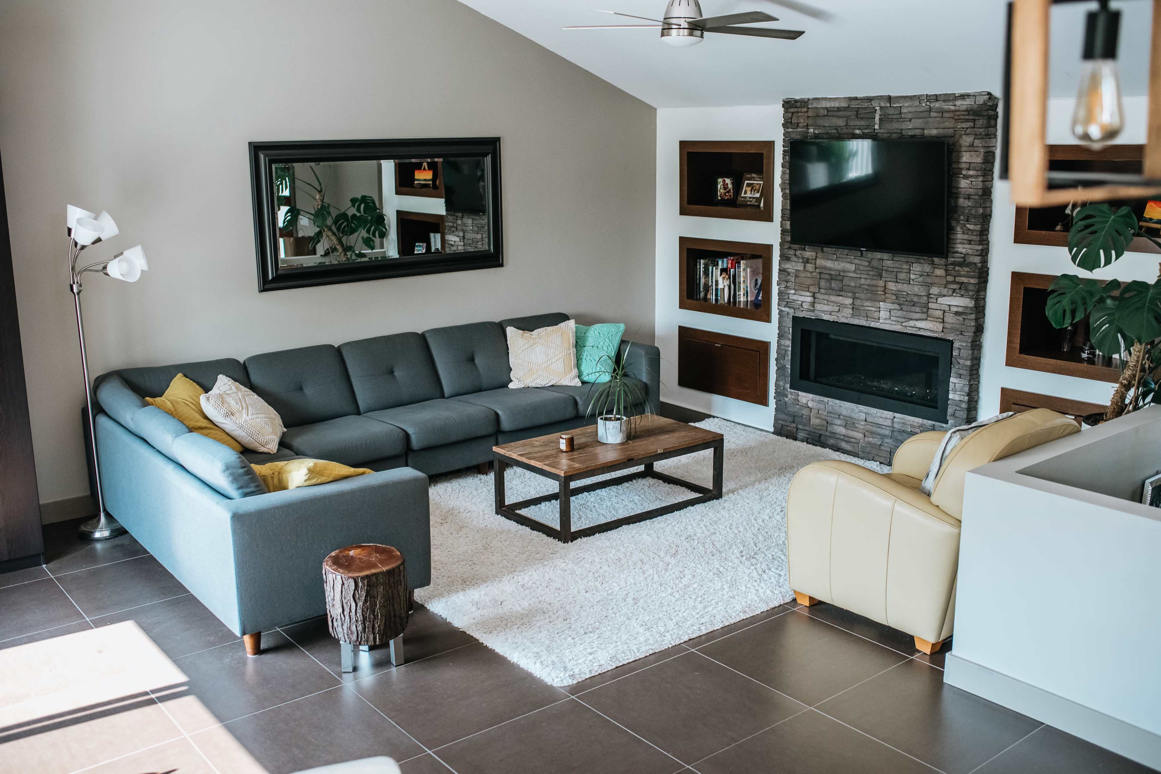A modern living room features a gray sectional sofa, a wooden coffee table, and a stone fireplace, all arranged on a light rug with decorative elements.