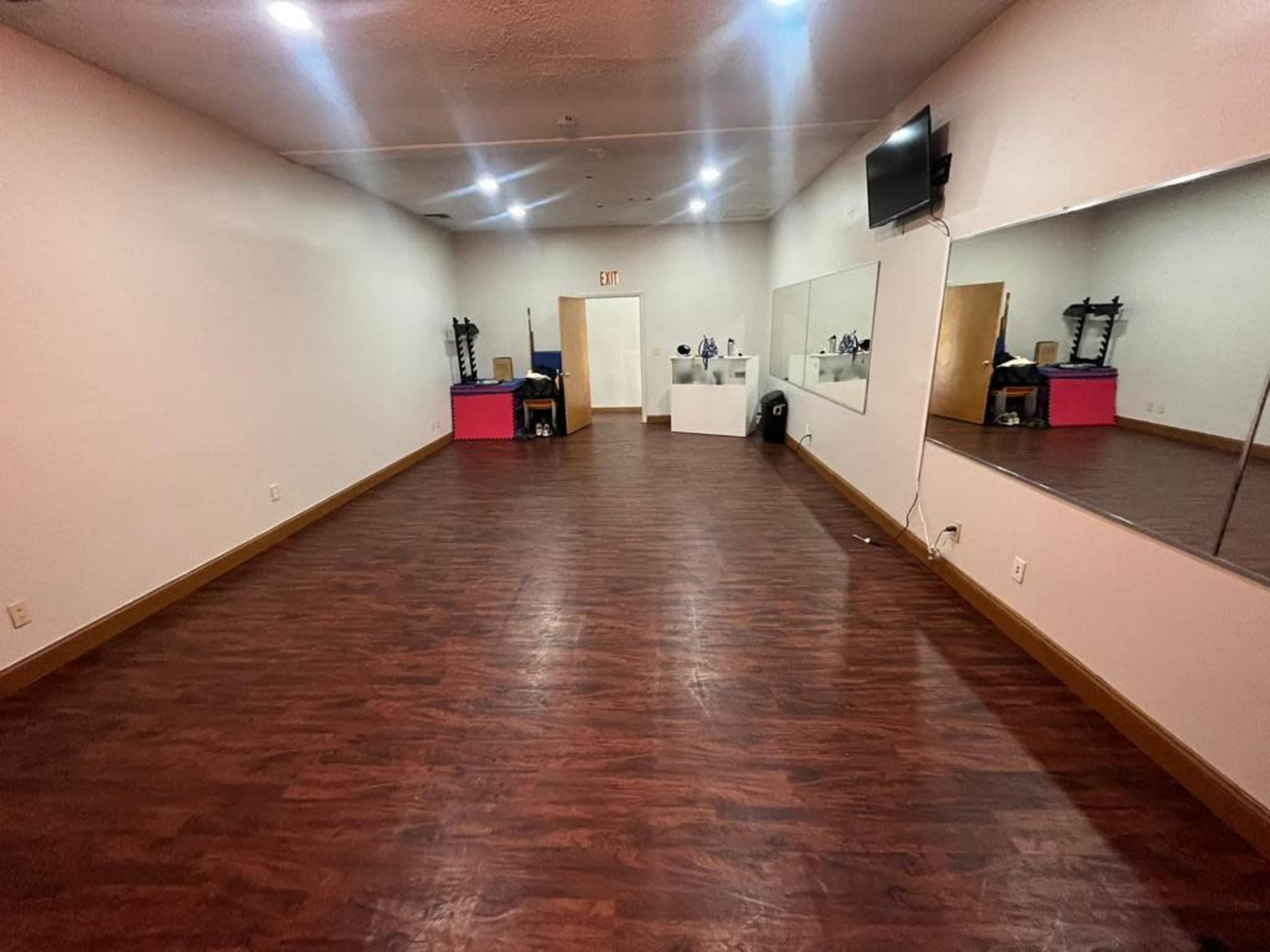 A spacious, empty room with wooden flooring, a large wall mirror, exercise equipment, and a wall-mounted television.