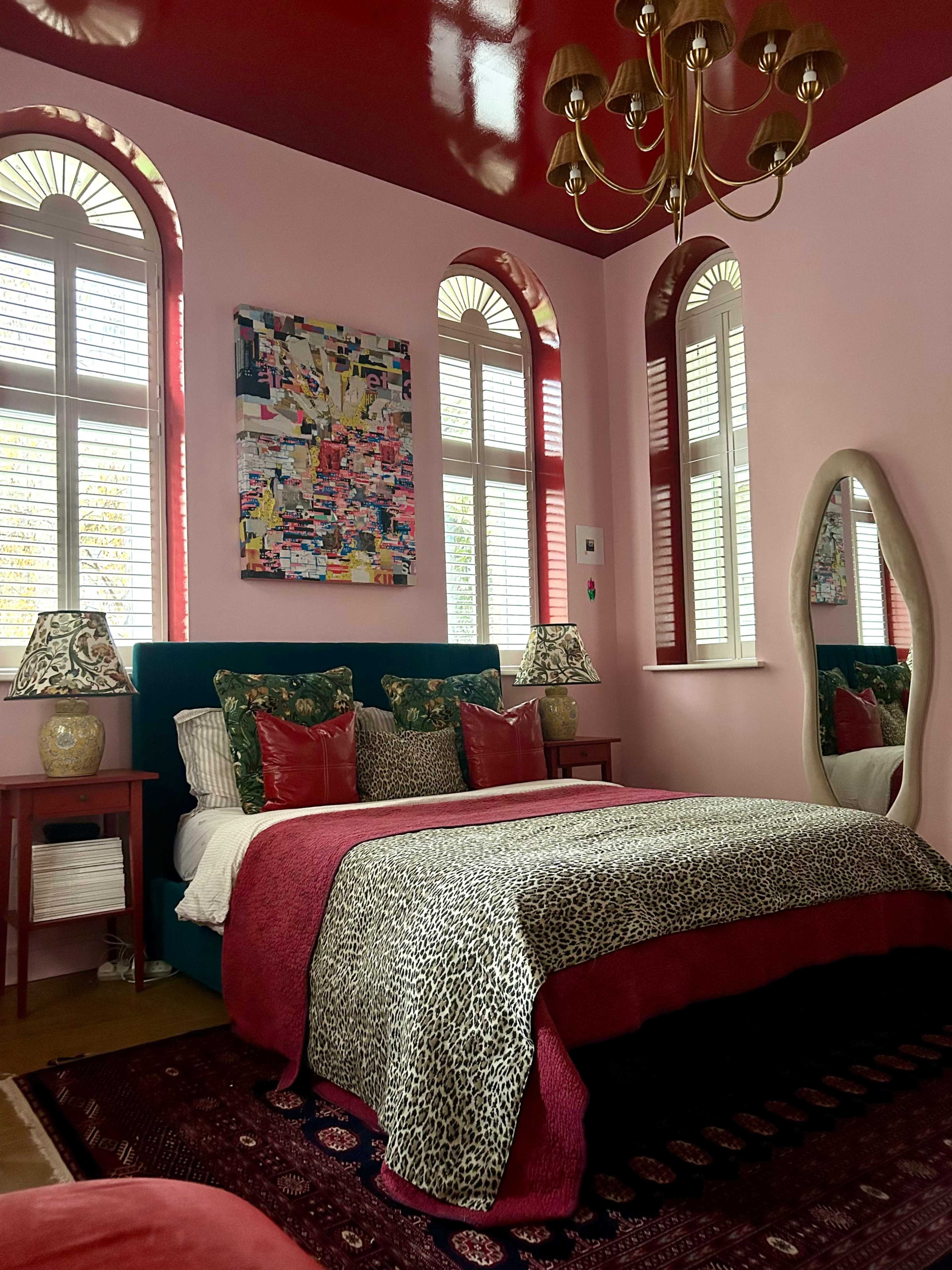 The image shows a stylish bedroom with a pink and red color scheme, featuring a large bed with patterned bedding, decorative lamps, and a framed artwork on the wall.