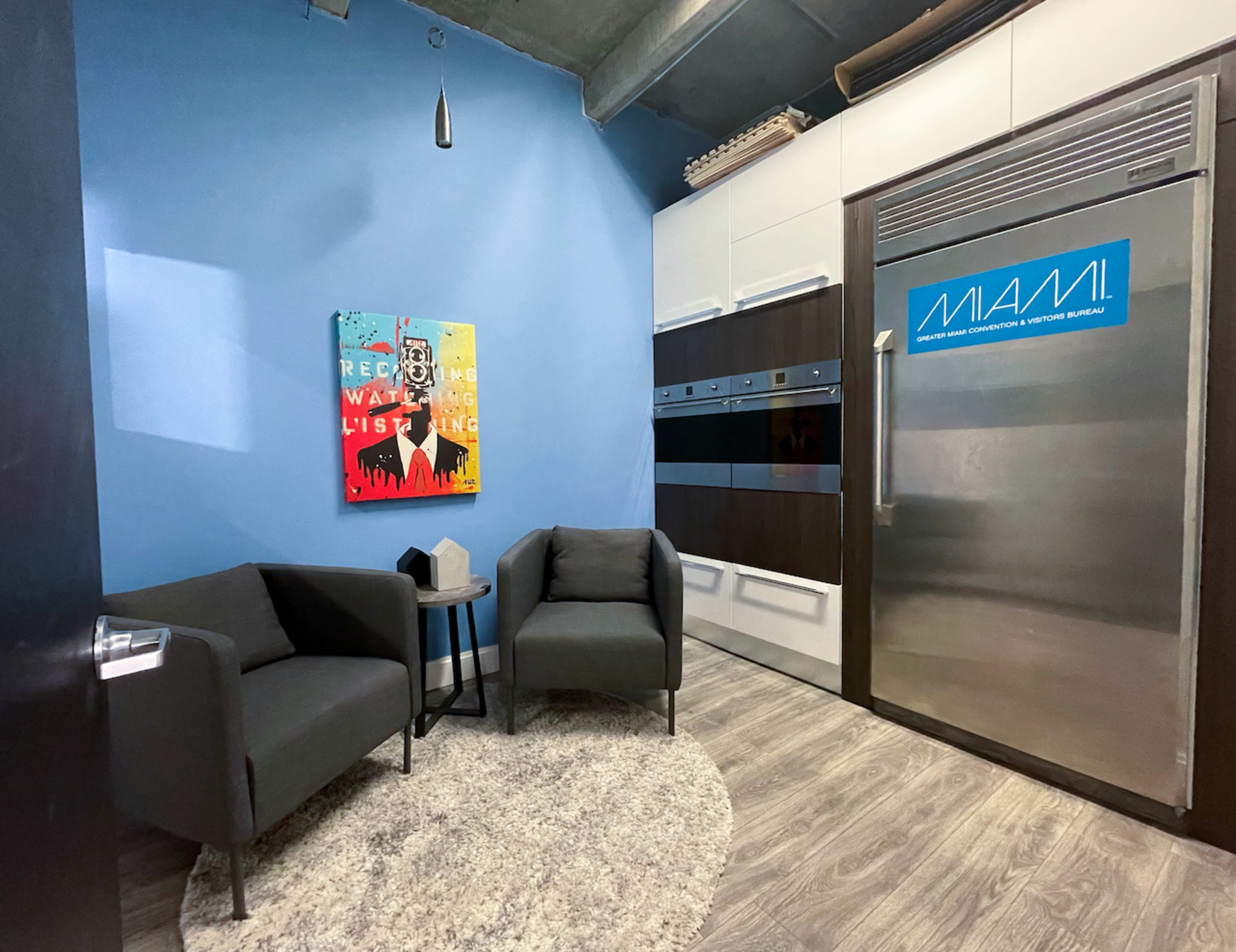 The image shows a modern interior with two gray armchairs, a small side table, a colorful artwork on the wall, and a stainless steel refrigerator labeled "MIAMI."
