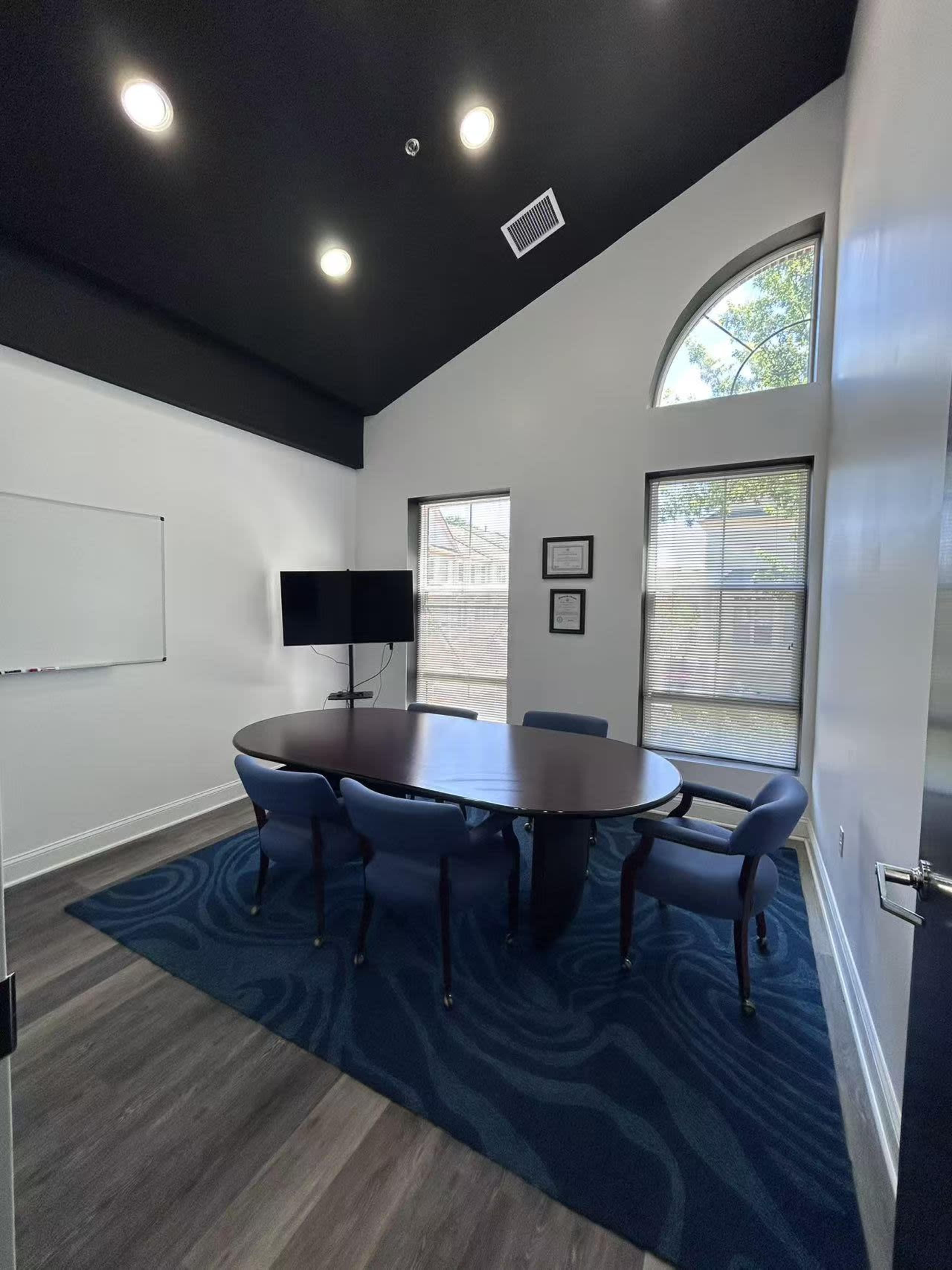 A conference room features a round table surrounded by blue chairs, a whiteboard, a television, and large windows with a view of the exterior.