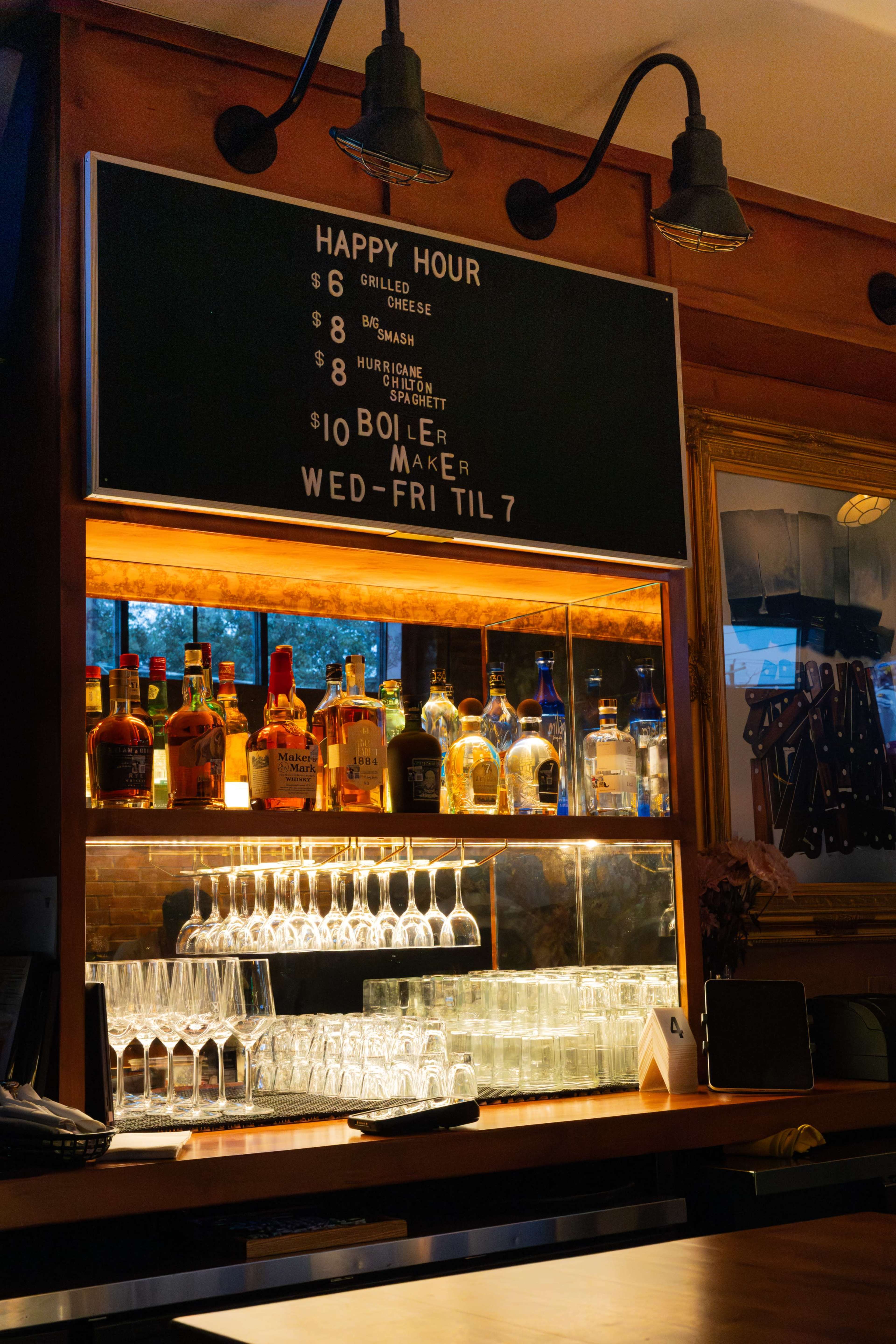 The image shows a bar counter with a menu board above displaying happy hour specials and a shelf stocked with various liquor bottles and glassware.