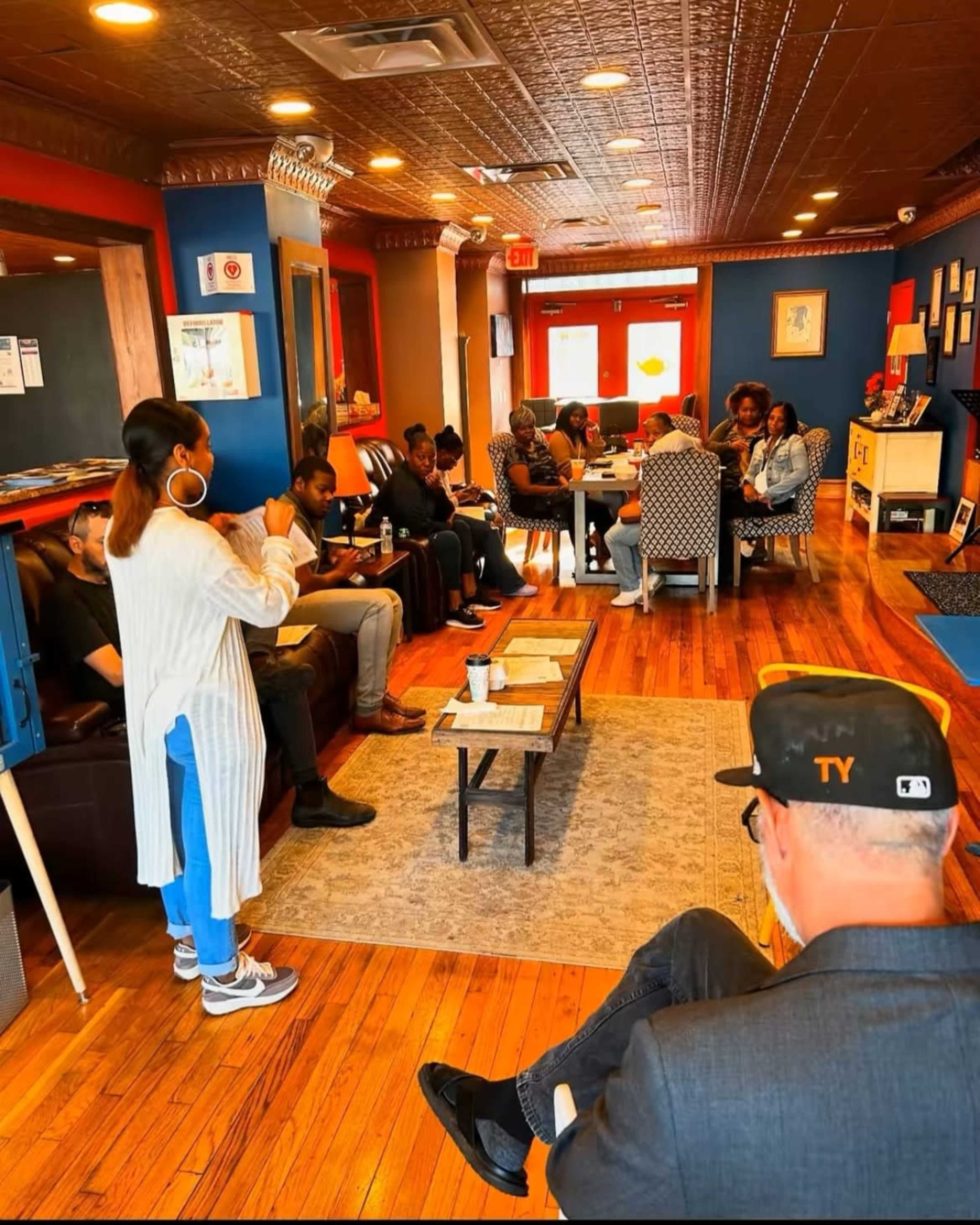 A group of individuals sit in a cozy, warmly-lit café while one person stands and speaks in front of them.
