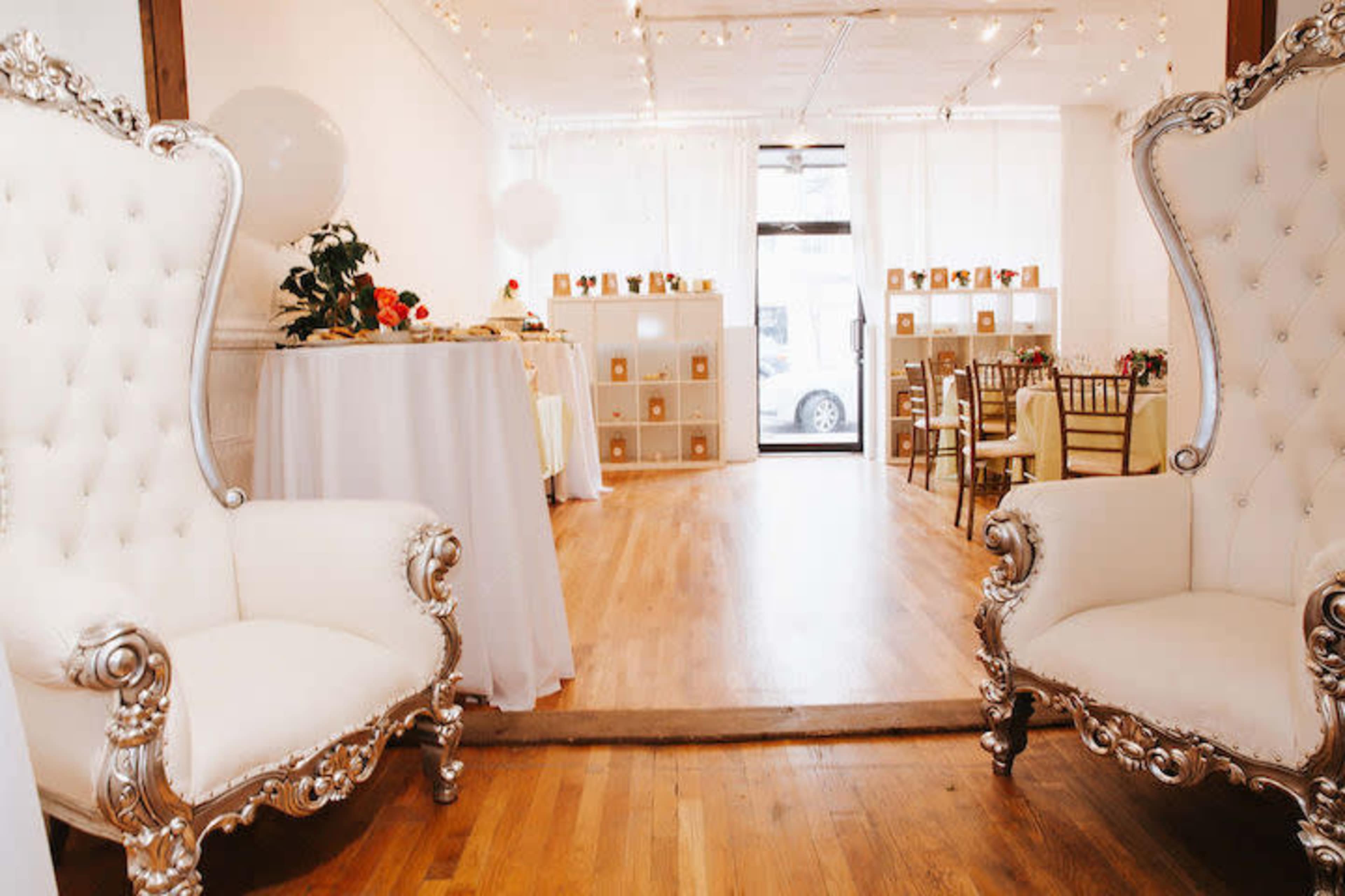 A spacious event room with two ornate white chairs at the front and a decorated table and seating area in the background.