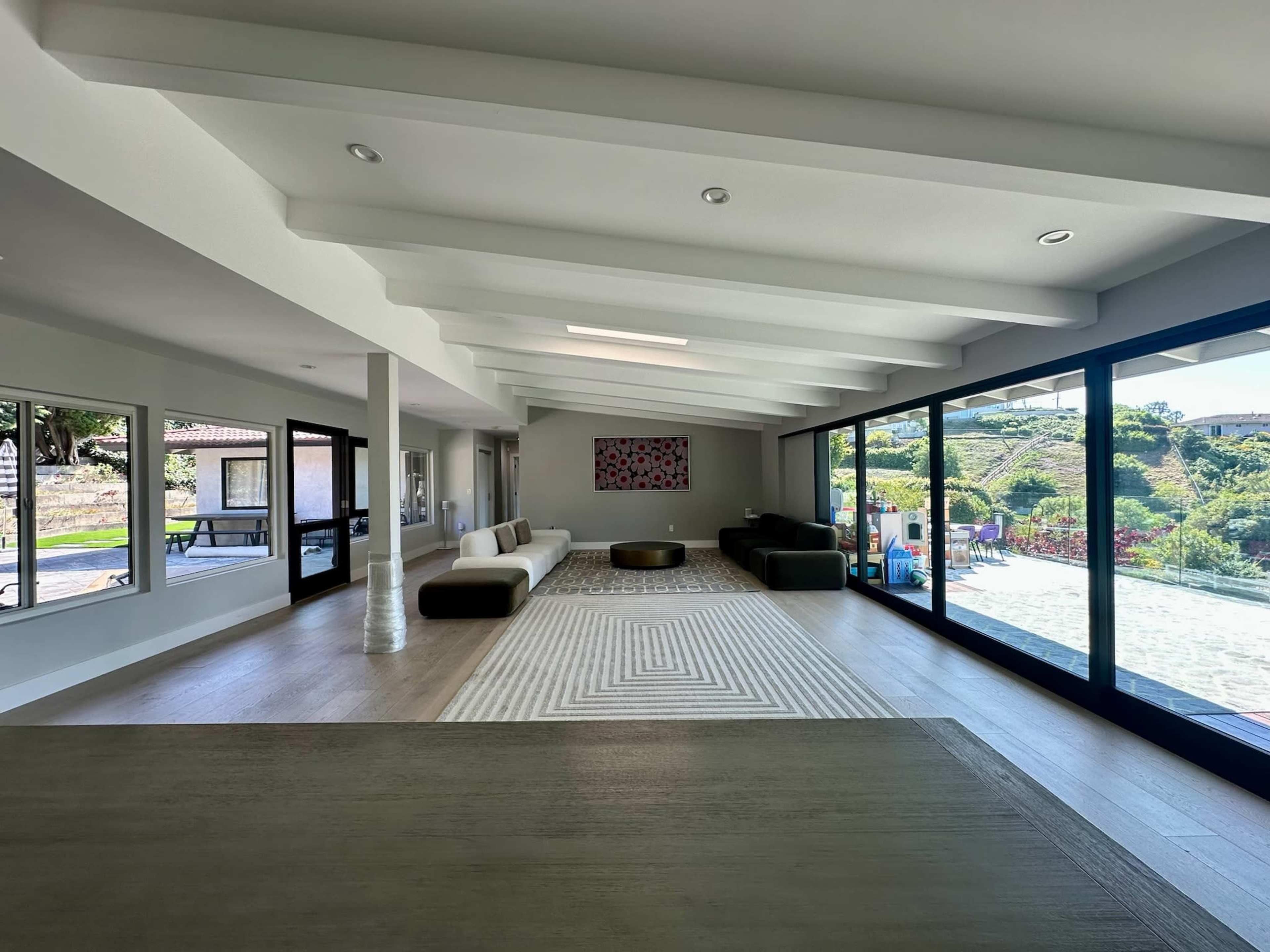 A spacious living room features large windows, a patterned rug, and modern furnishings overlooking a landscaped exterior.