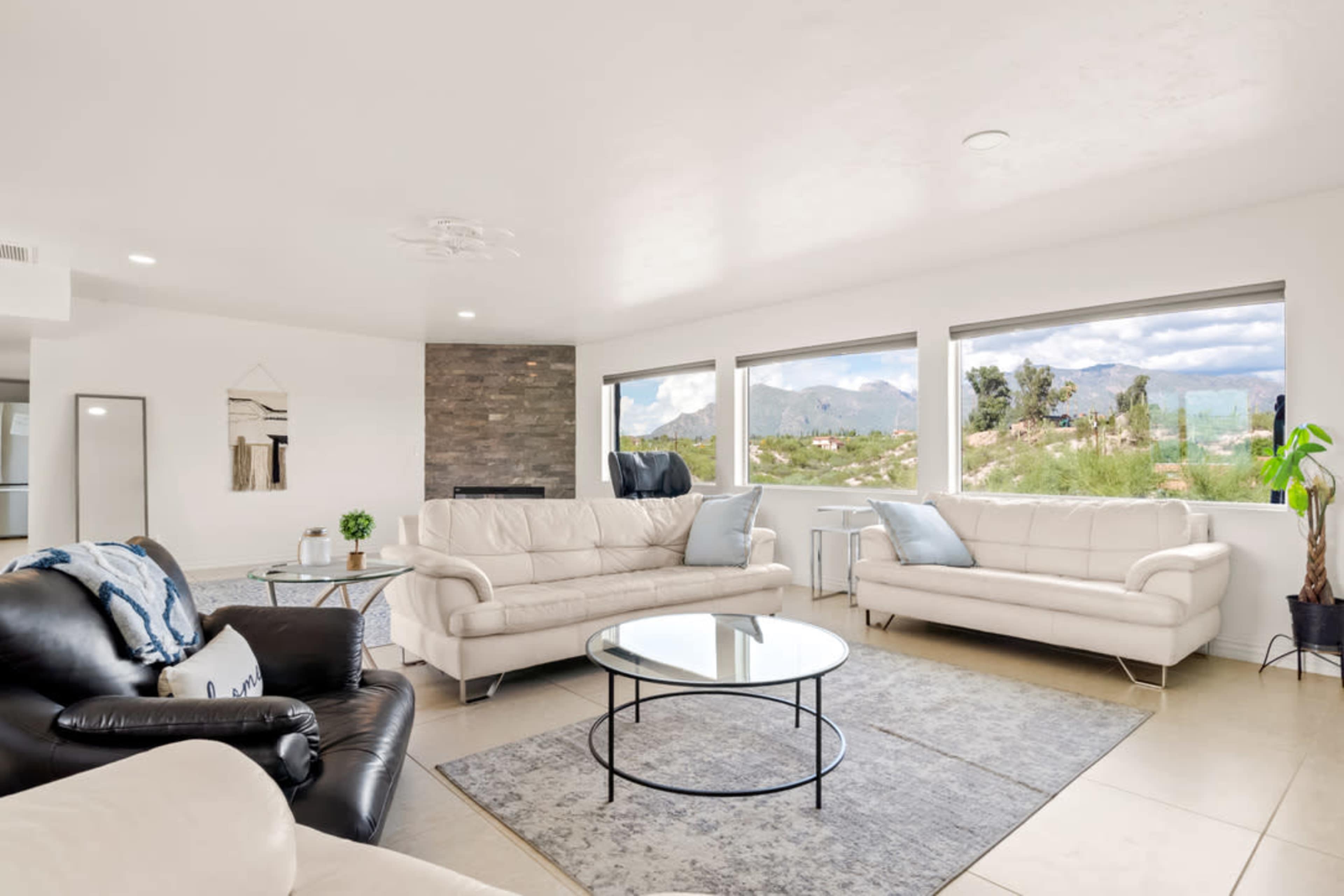 The living room features two light-colored sofas and one dark leather chair arranged around a glass coffee table, with large windows offering a view of the mountains.