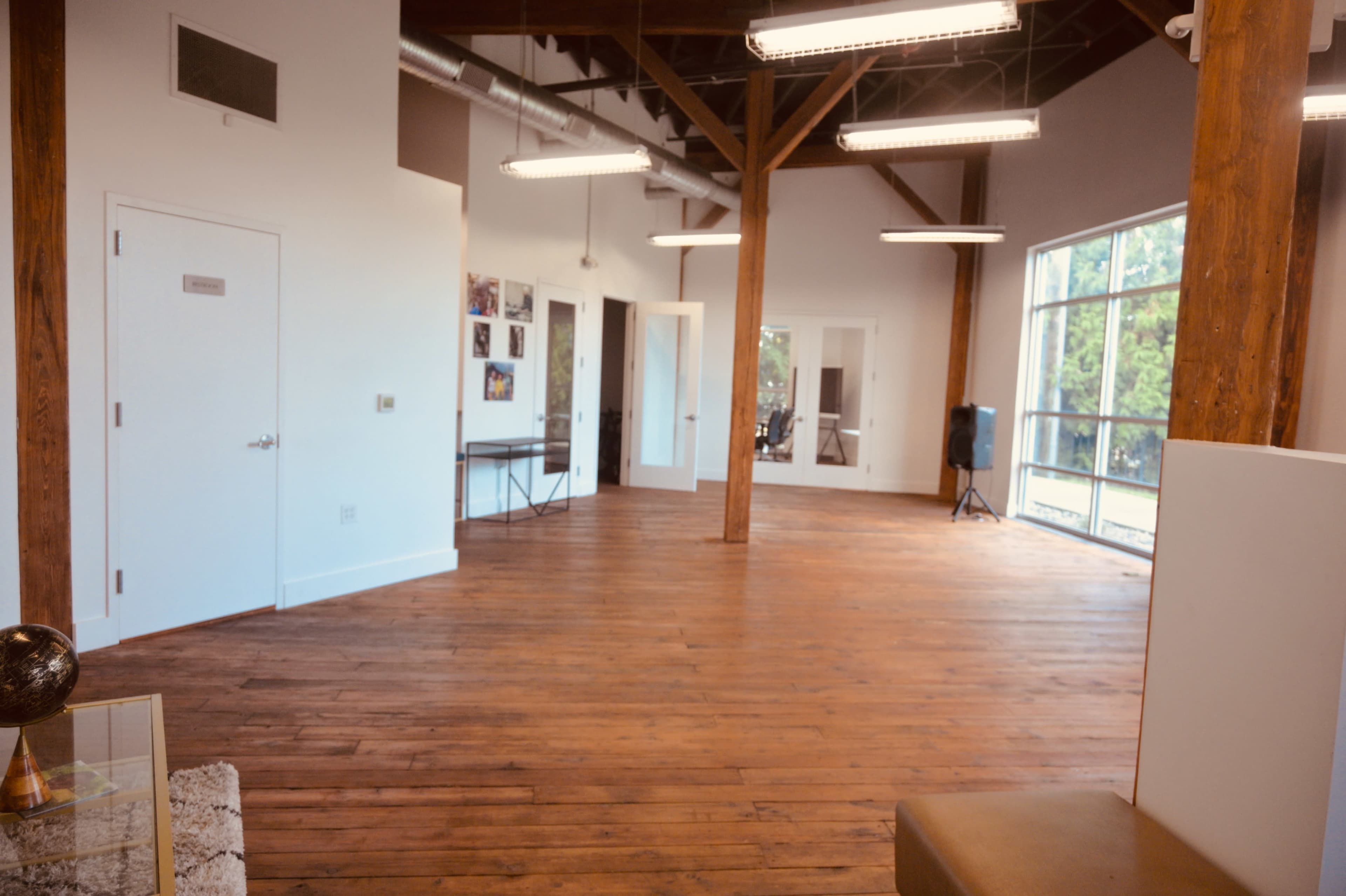 The image shows a spacious interior with wooden floors, tall ceilings supported by wooden beams, and large windows that provide natural light.