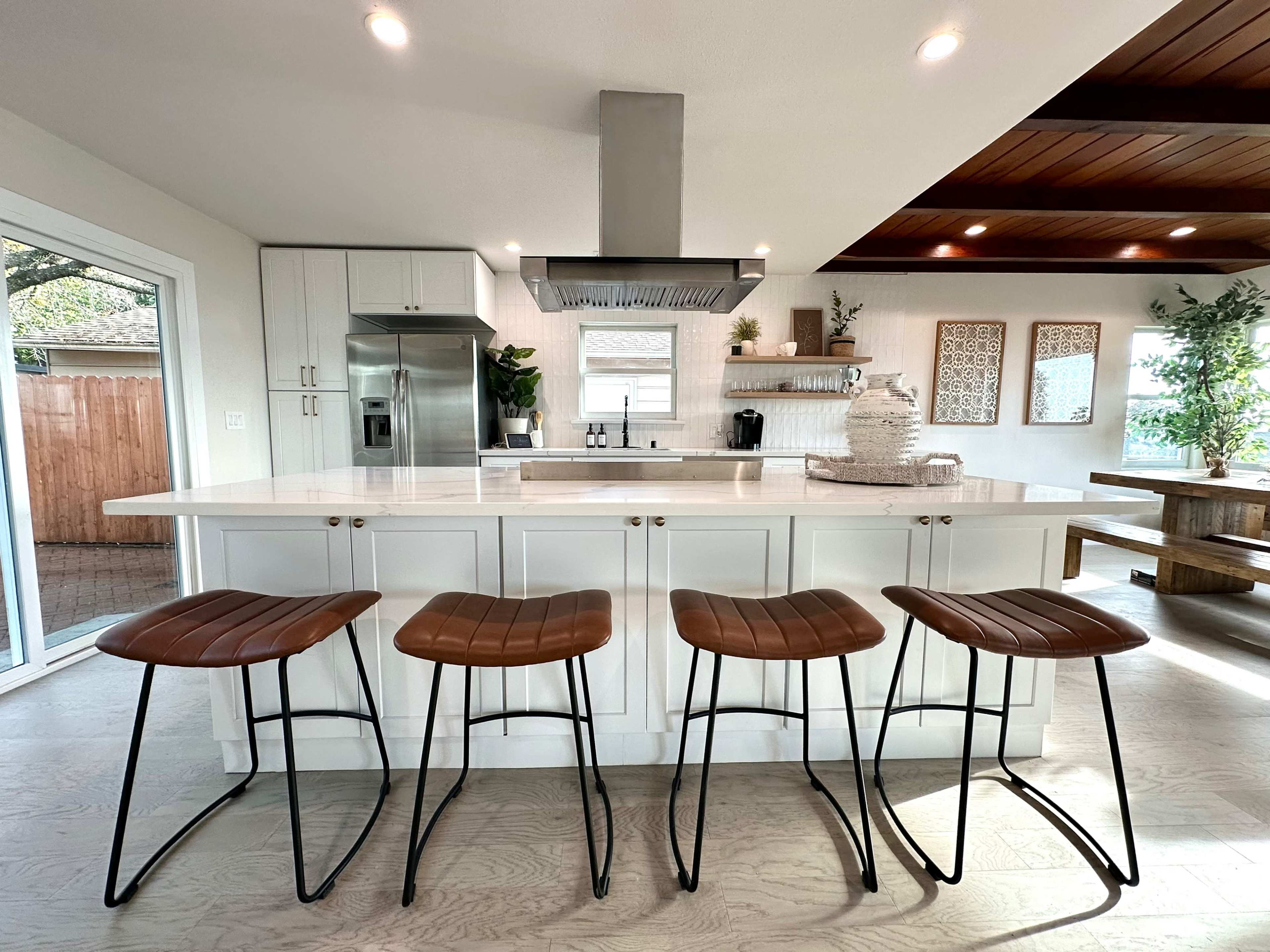 A modern kitchen with a central island featuring four metal-framed, wooden seating stools and stainless steel appliances.