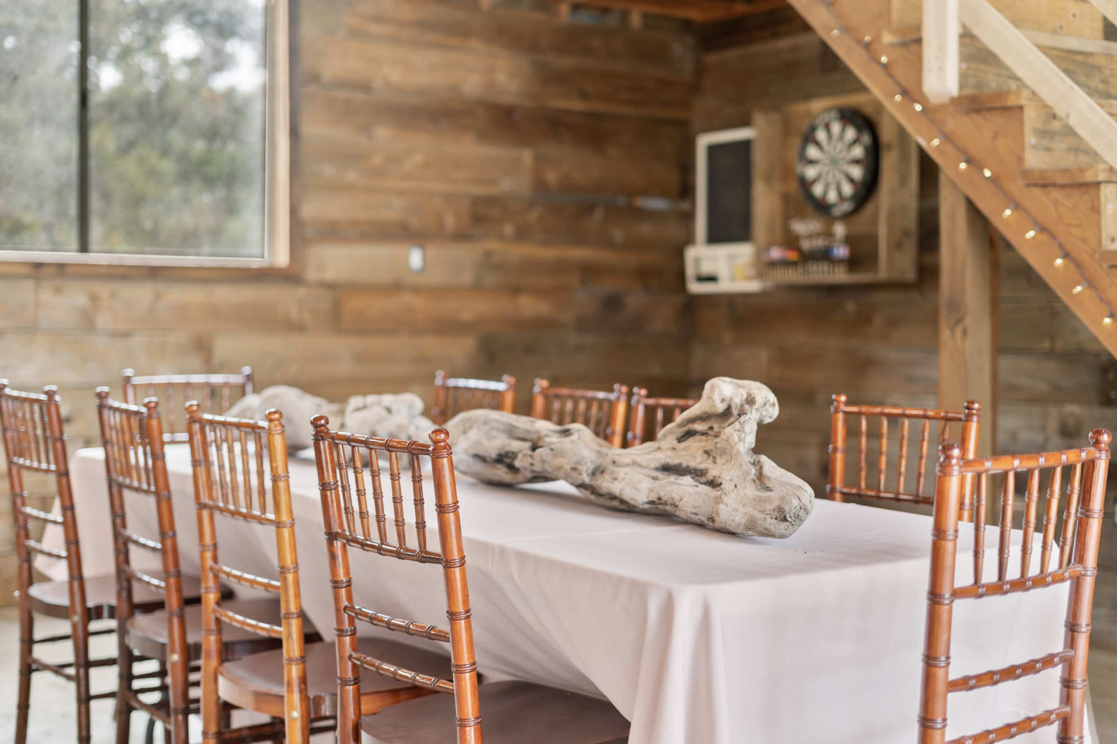 A long table with a white tablecloth is set in a wooden room, surrounded by multiple wooden chairs, and features a decorative piece of driftwood at the center.
