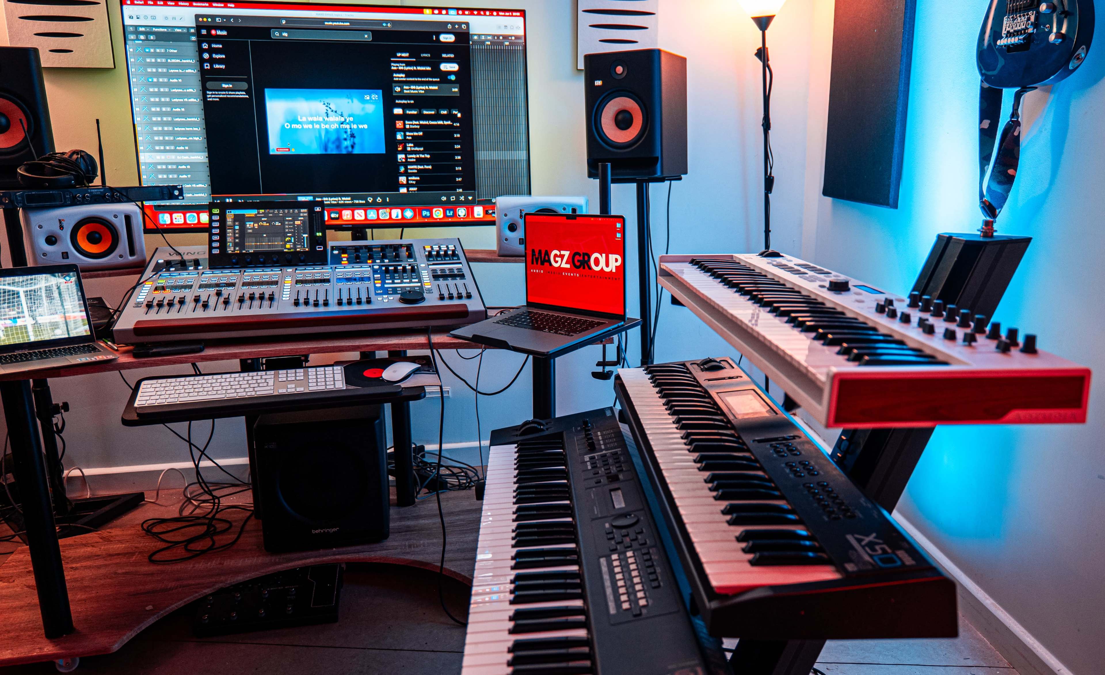 The image shows a music production studio featuring multiple electronic instruments, a mixing console, a laptop, and a large monitor displaying software.