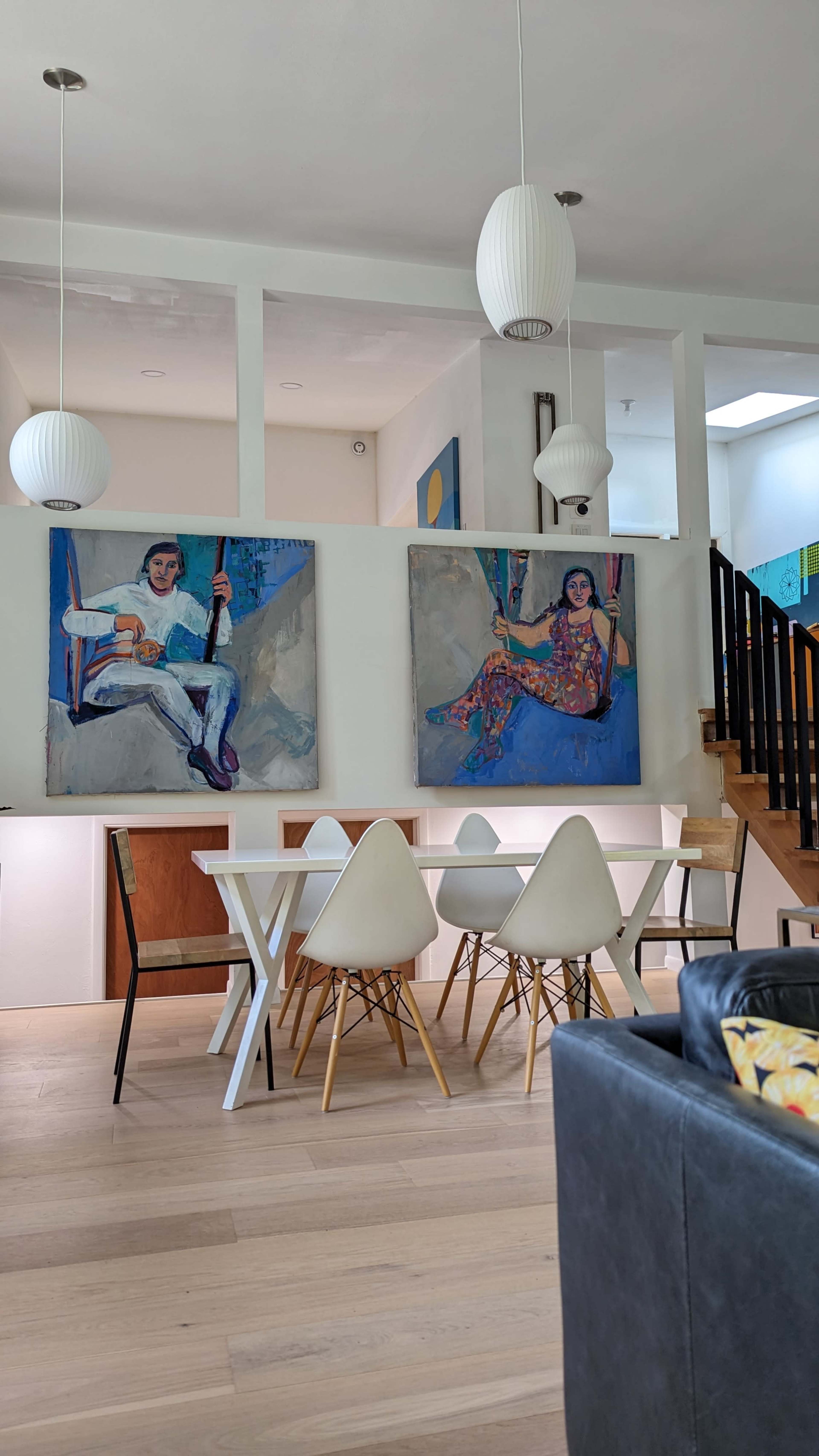 The image features a modern dining area with a white table and chairs, two large paintings on the walls, and a staircase in the background.