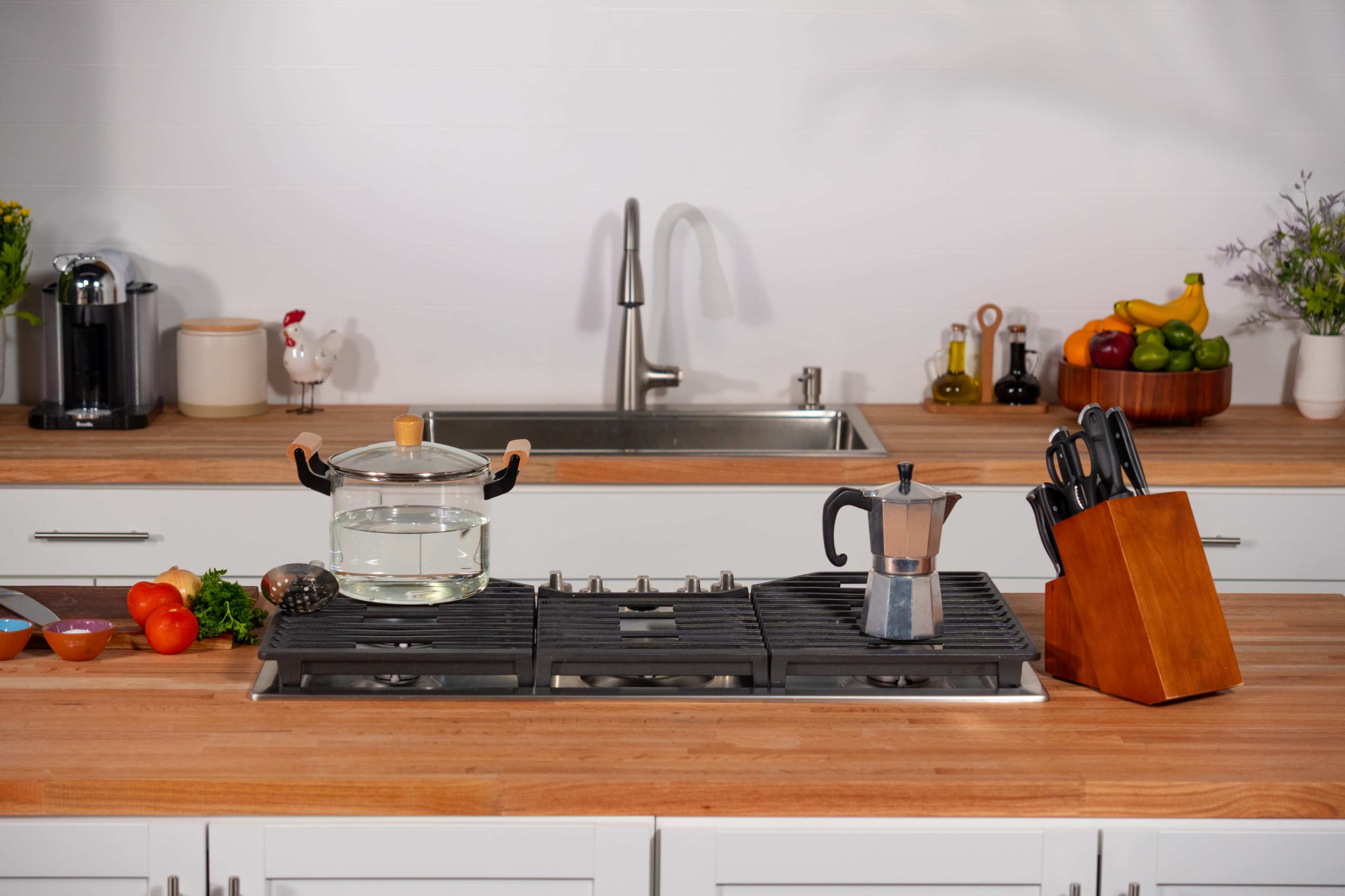 A modern kitchen countertop features a glass pot with water, a coffee maker, fresh vegetables, and a knife block, with a sink in the background.