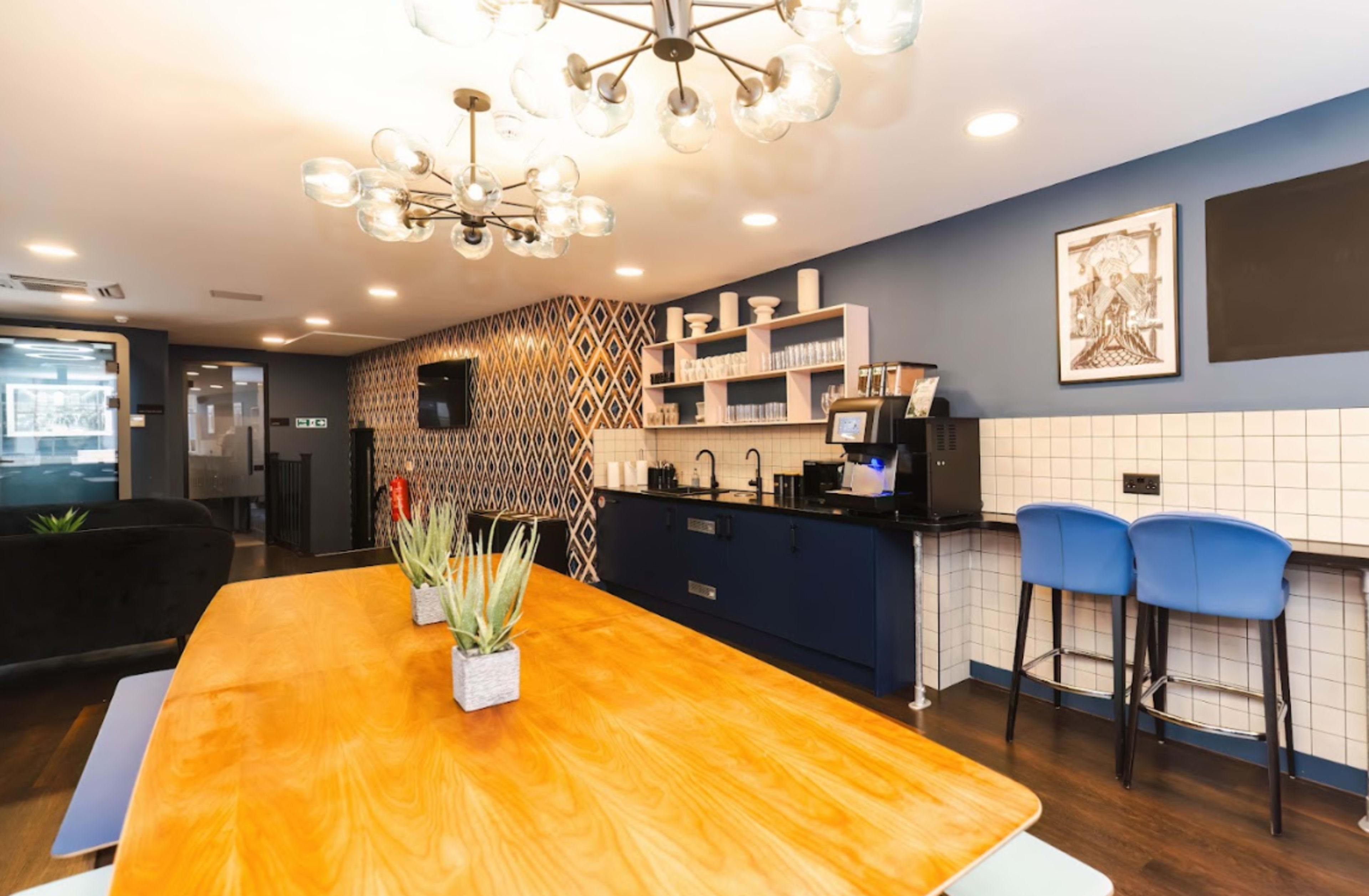 A modern interior space with a wooden dining table, blue chairs, and a kitchen area featuring a coffee machine, glassware, and decorative wall patterns.