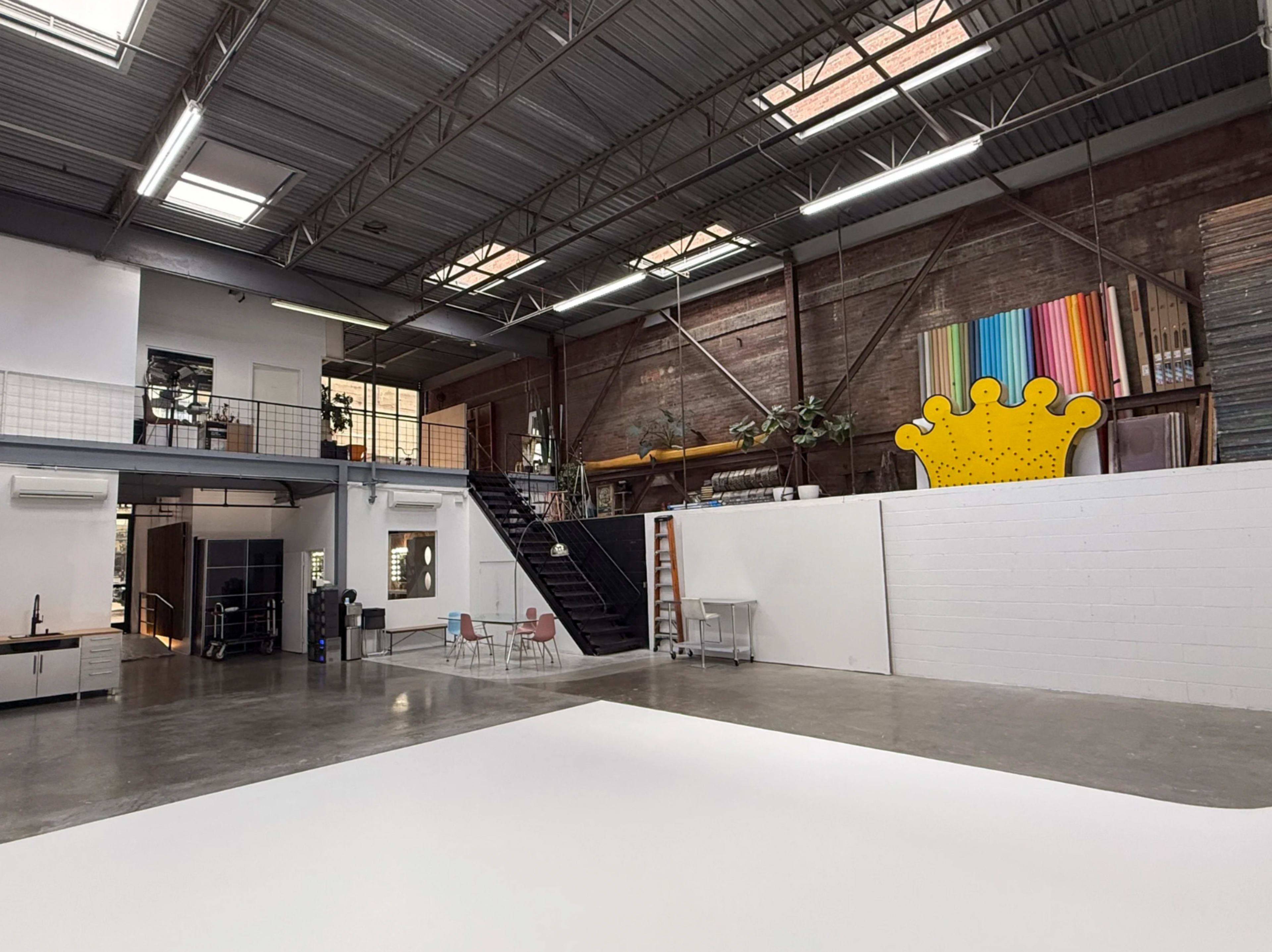 The image shows a spacious industrial-style studio with high ceilings, large windows, and a loft area containing colorful artwork.
