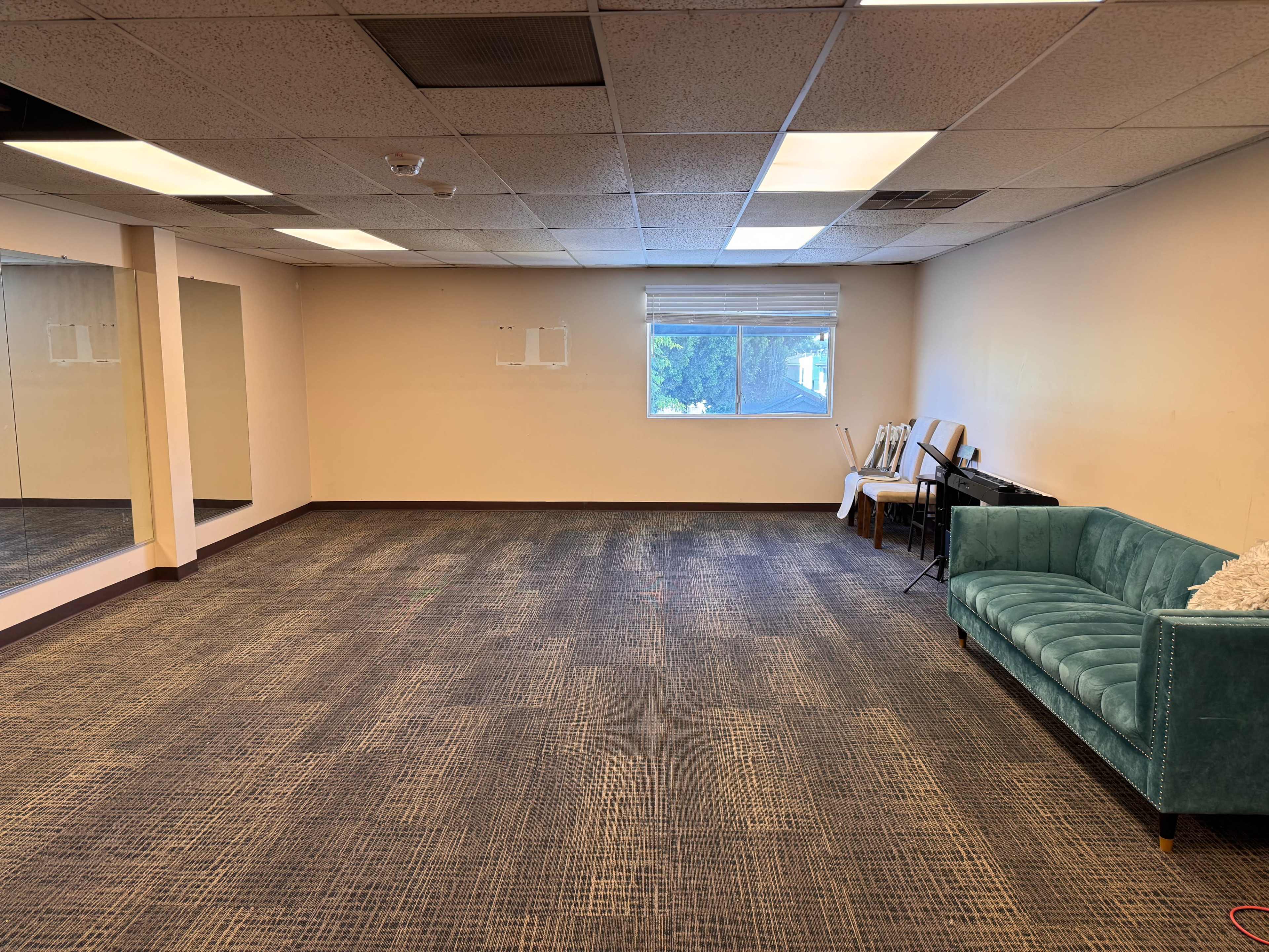 The image shows a spacious room with a large mirror on one wall, a window letting in natural light, a teal couch, and a few chairs and a keyboard against the opposite wall.