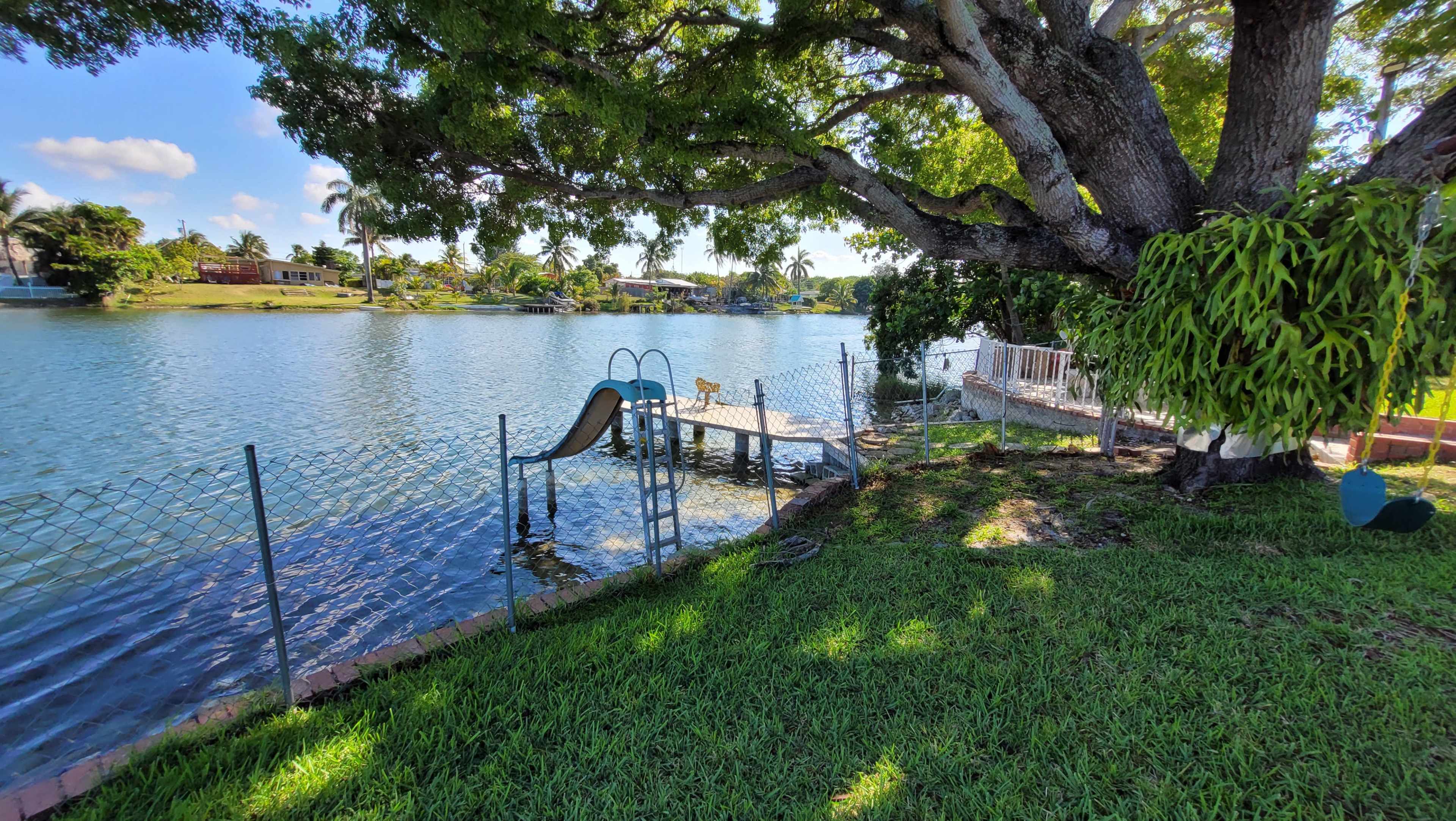 A grassy area with a waterslide leads to a wooden dock by a calm lake, surrounded by trees and a fence.