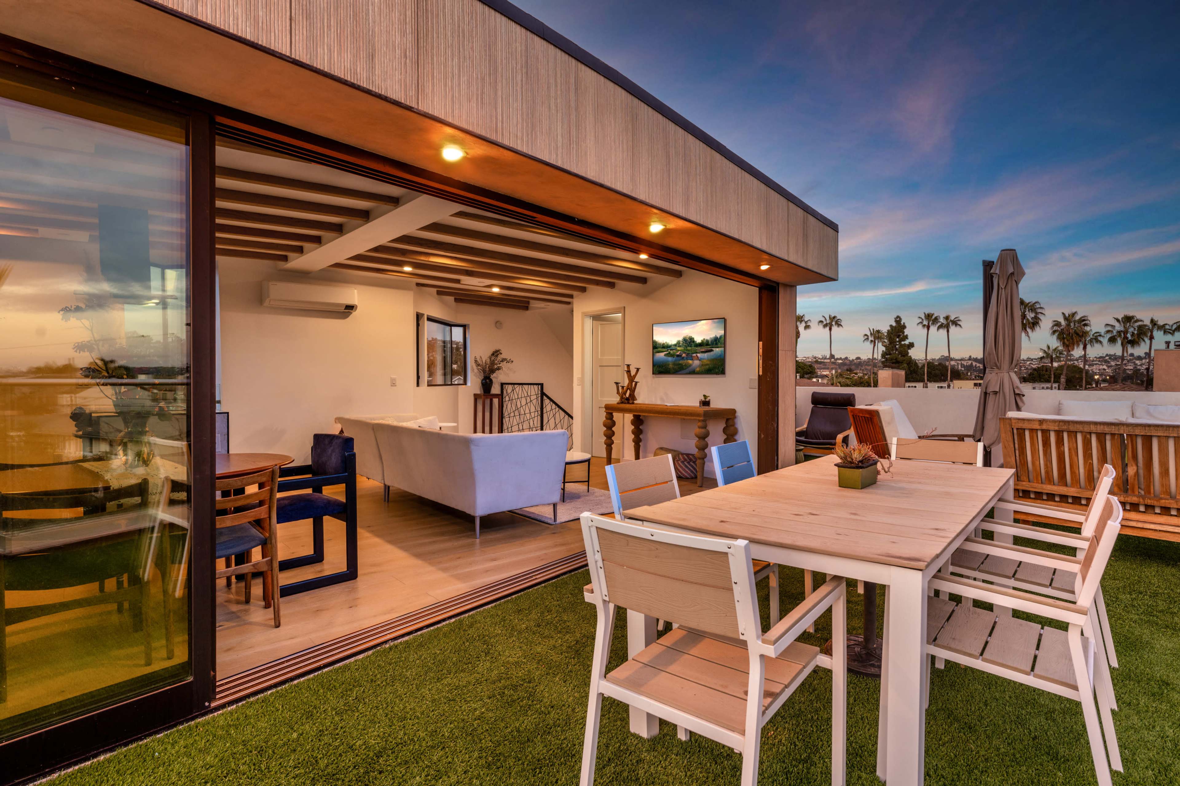 A modern rooftop terrace features a dining table surrounded by white chairs, overlooking a sunset with palm trees in the distance.