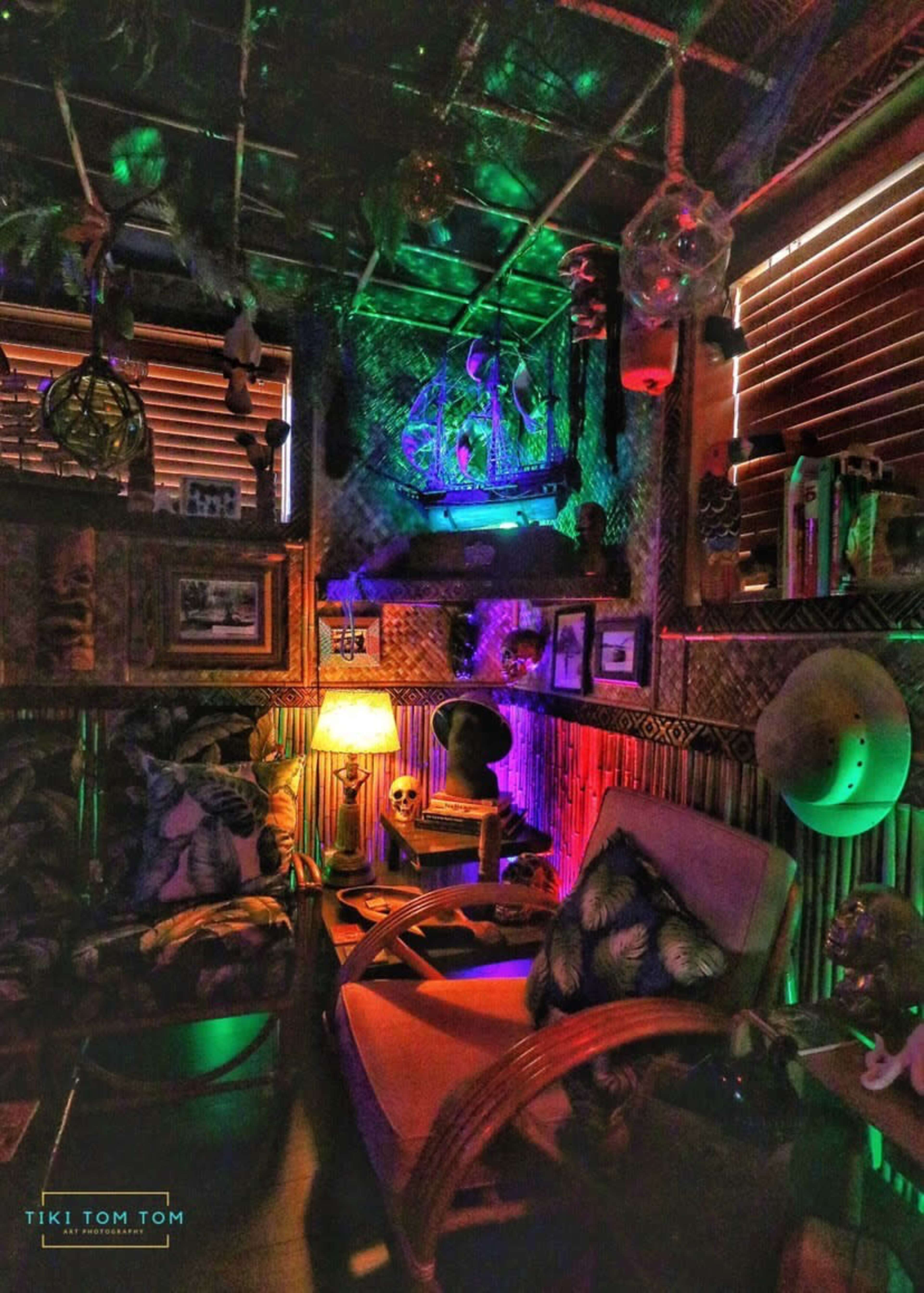 A dimly lit, eclectic tiki-themed room filled with tropical decor, colorful lighting, and various decorative objects.