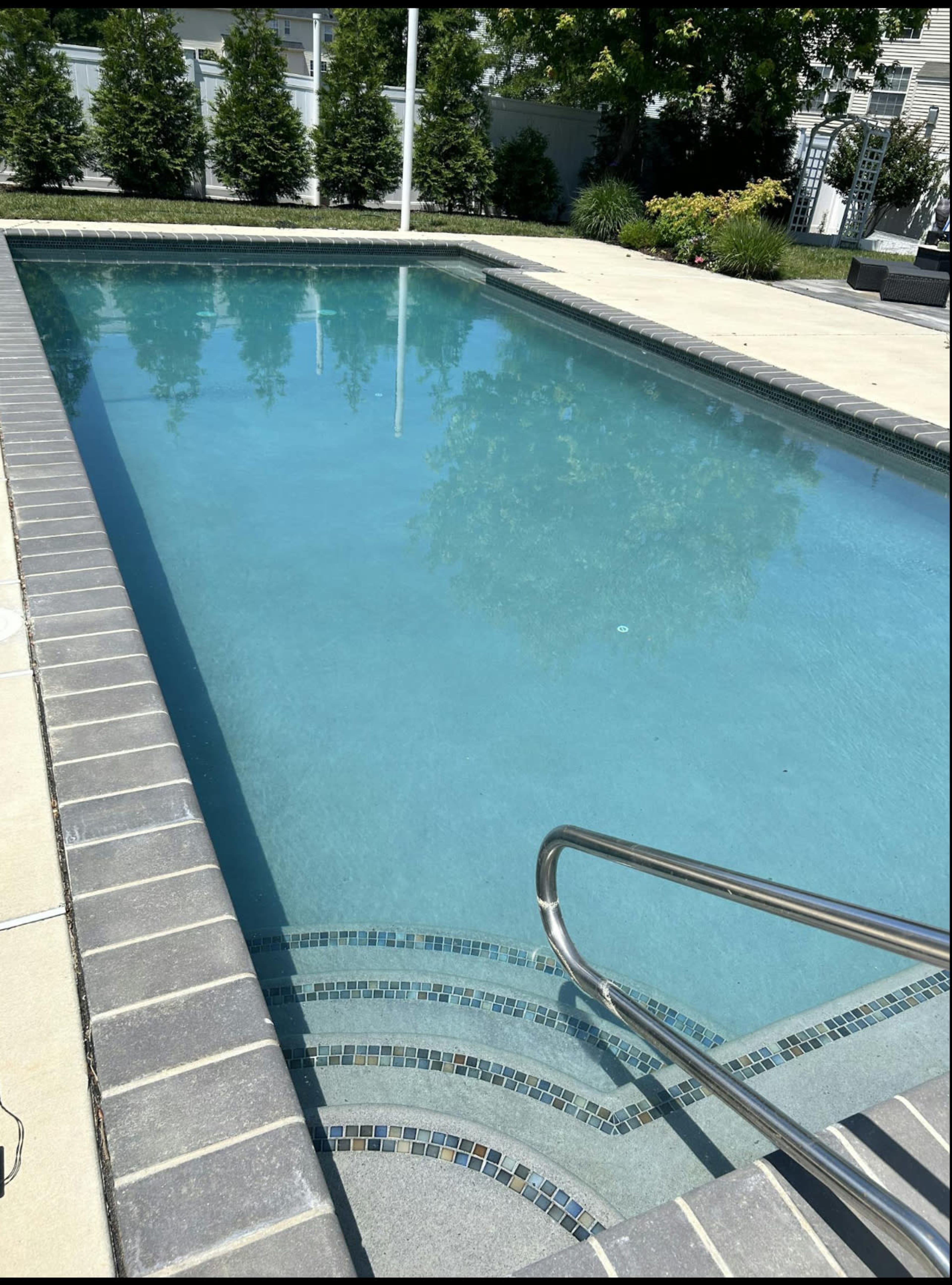 The image shows a clear swimming pool with a set of metal stairs leading into it, surrounded by a tiled deck and landscaped greenery.