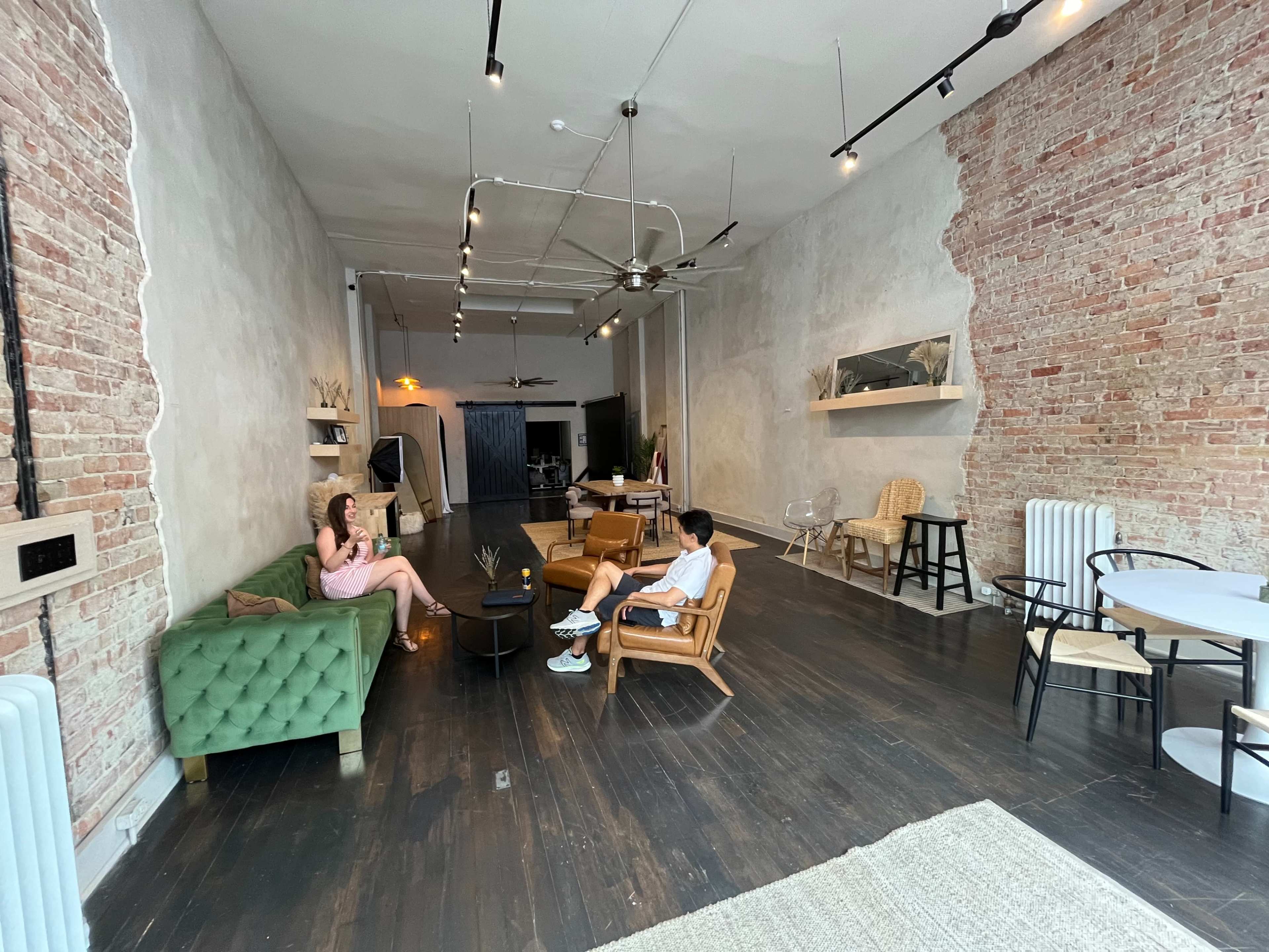 The interior of a modern, industrial-style room features a green velvet sofa, wooden chairs, and exposed brick walls, with two people seated and engaged in conversation.