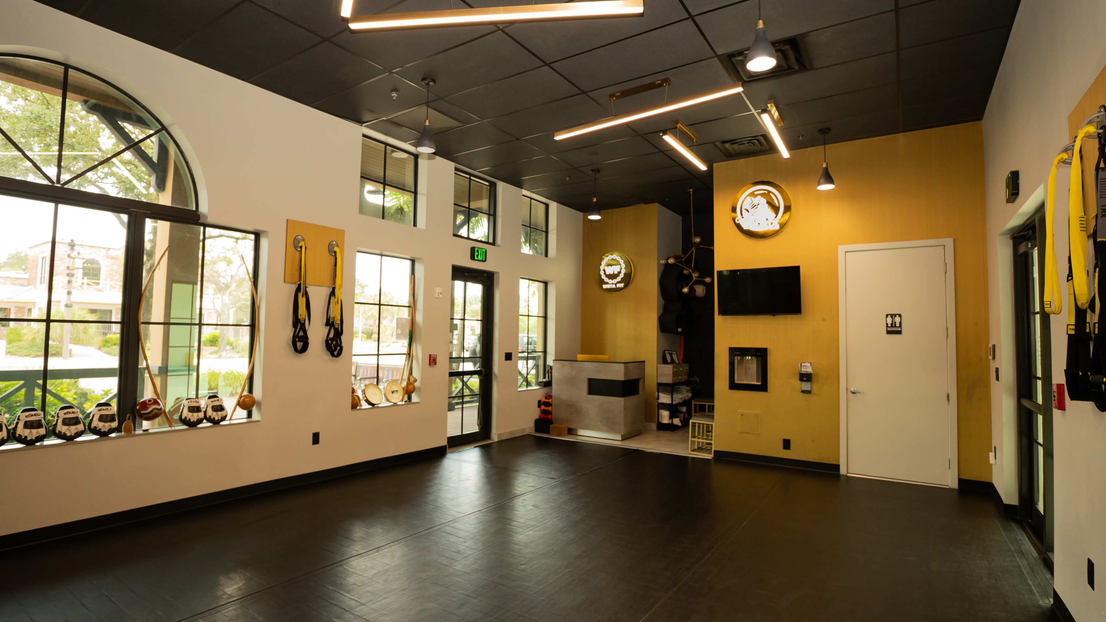 The image shows a fitness studio interior with large windows, black flooring, and exercise equipment such as TRX straps and kettlebells displayed on the walls.