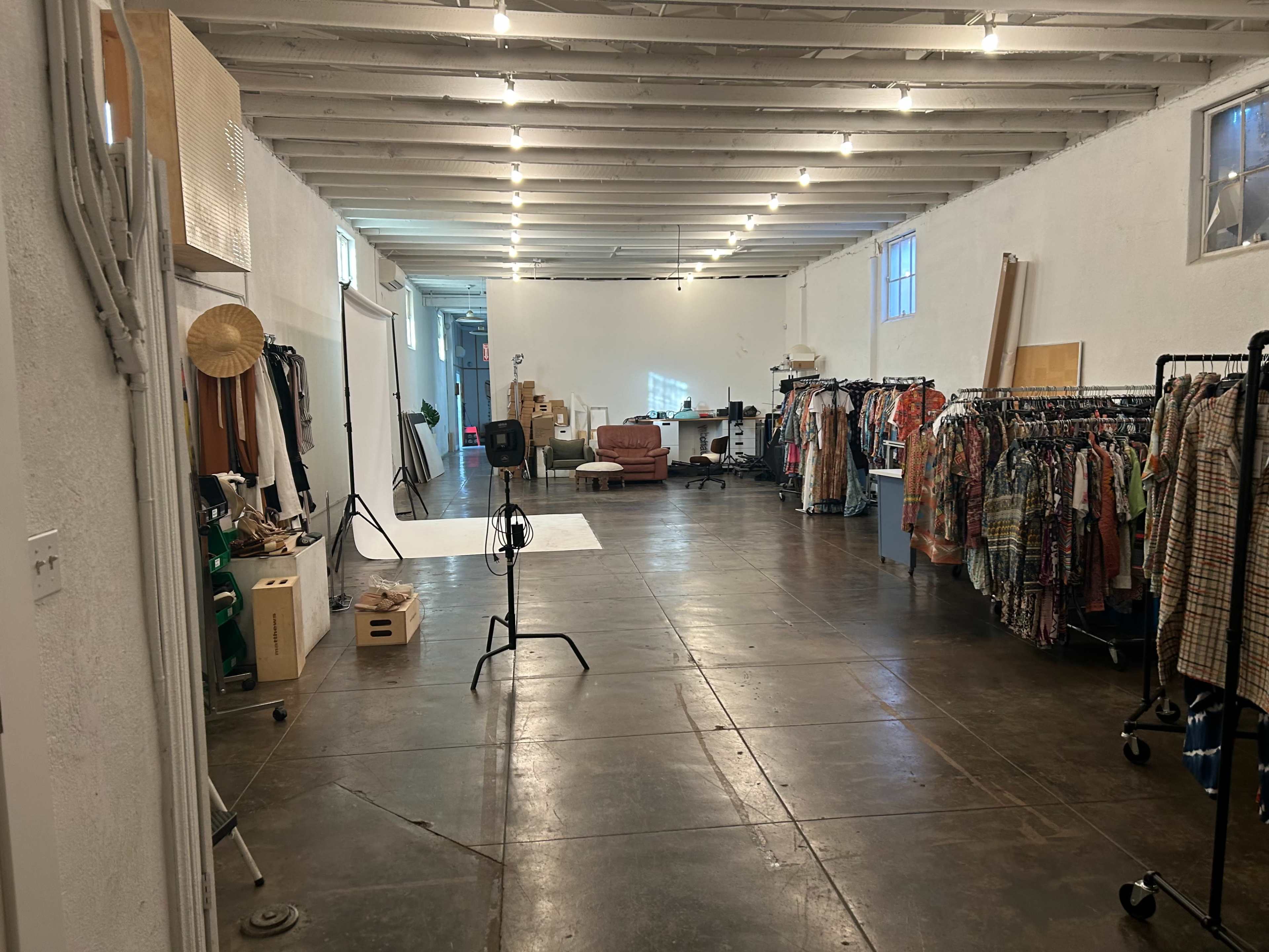 A spacious room with clothing racks filled with various garments, a white backdrop set up for photography, and a few pieces of furniture against the walls.