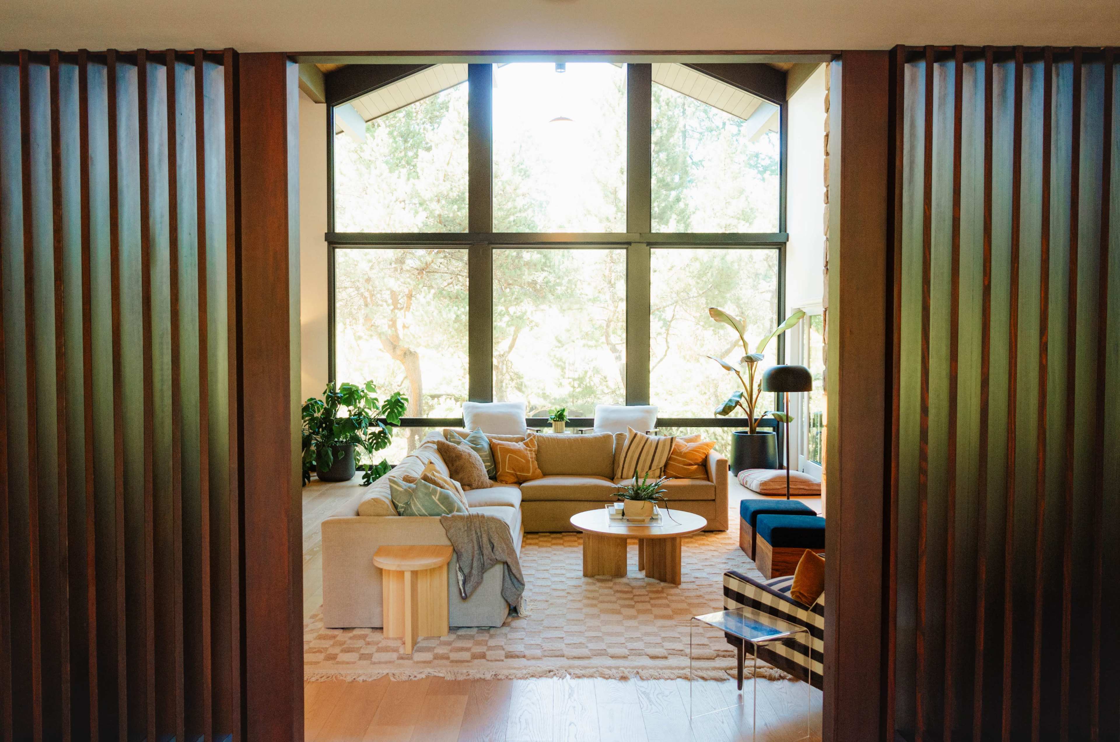A modern living room with large windows, a comfortable sofa, a round coffee table, and various plants in a sunlit setting.